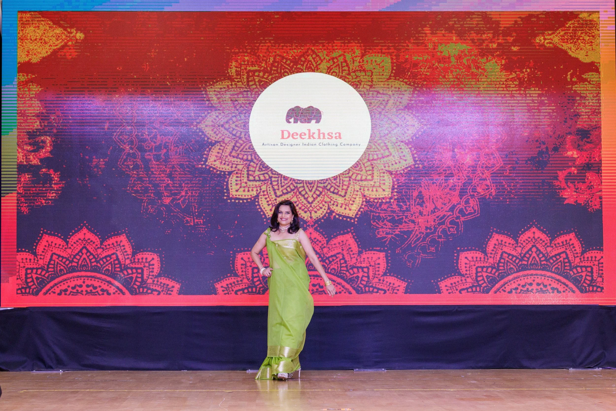 Woman in a lime-green saree posing on stage in front of a decorative backdrop with the logo 'Deeksha' and stylized patterns.
