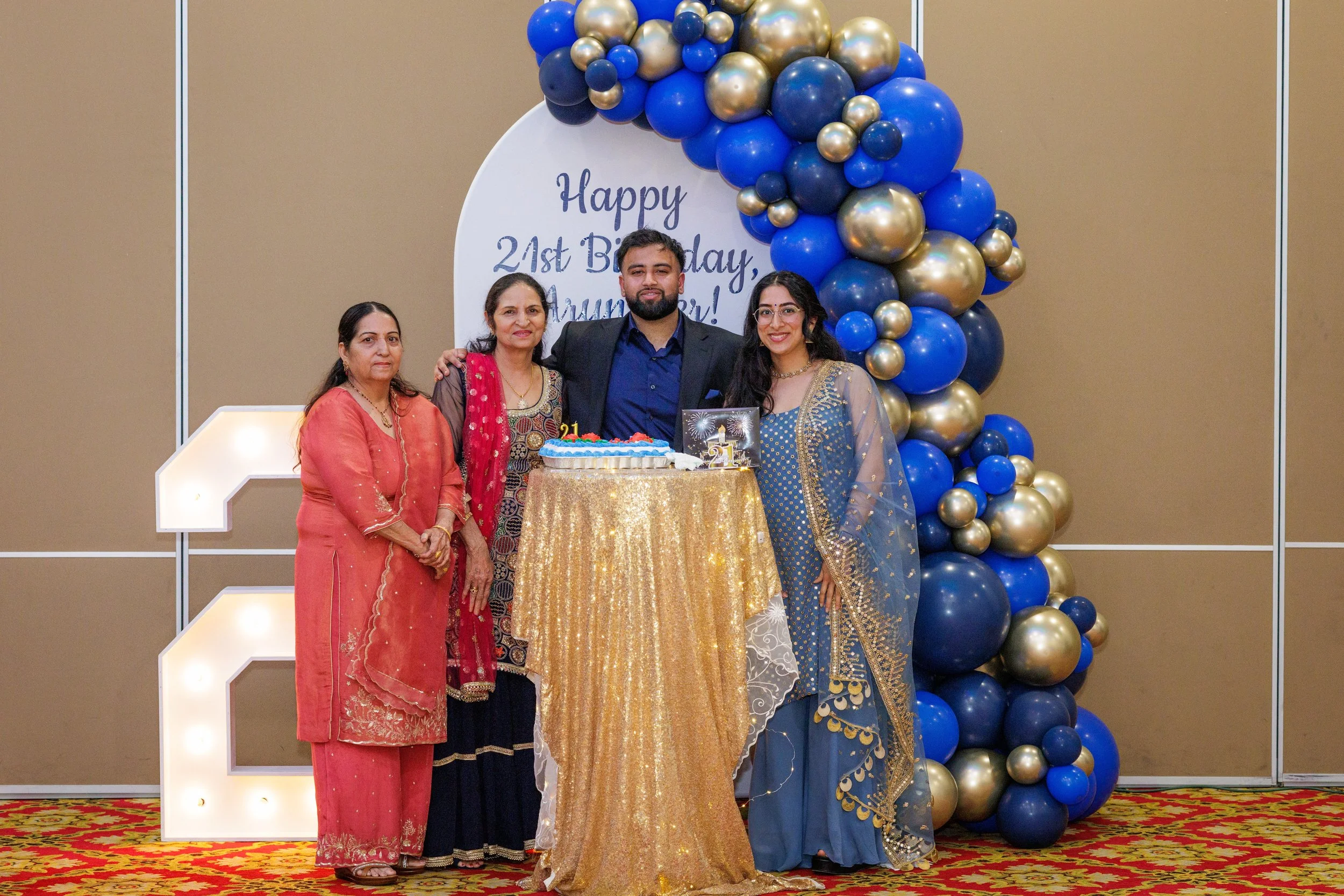 A family celebrating a birthday, posing behind a table with a cake. The backdrop has balloons in blue, gold, and black, and a sign that reads "Happy 21st Birthday". They are dressed in colorful traditional Indian clothing, with a decorated carpeted f