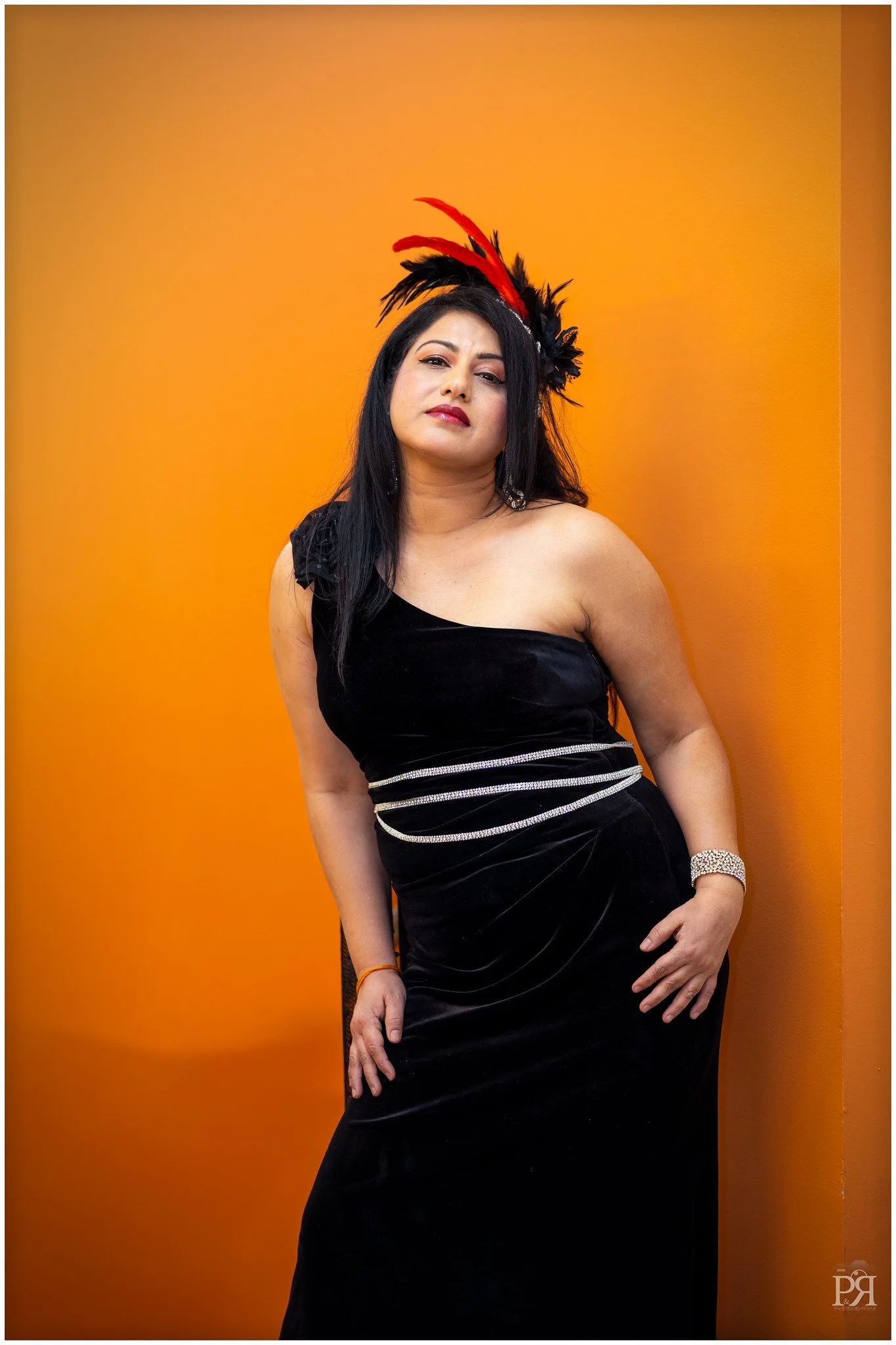 A woman with long black hair wearing a black strapless dress with rhinestone details, a feathered red and black headpiece, and a silver bracelet, standing against an orange background.