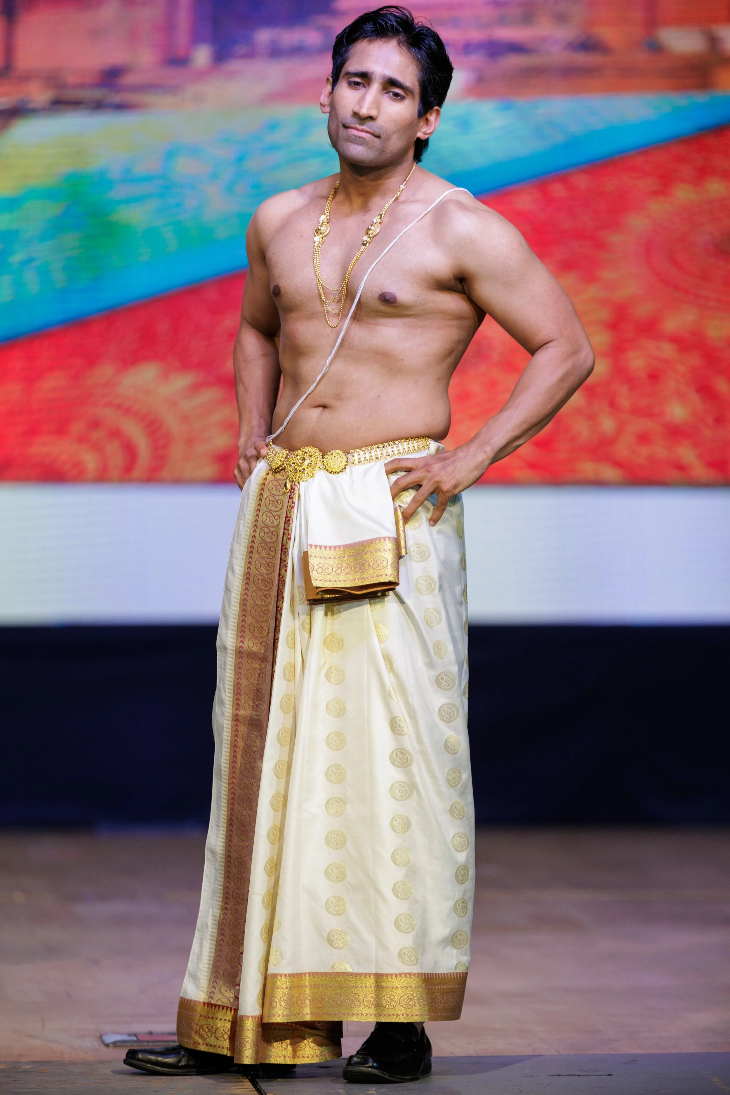 A man dressed in traditional Indian attire, wearing a gold dhoti with intricate patterns, gold jewelry, and posing confidently on stage with a colorful blurred background.