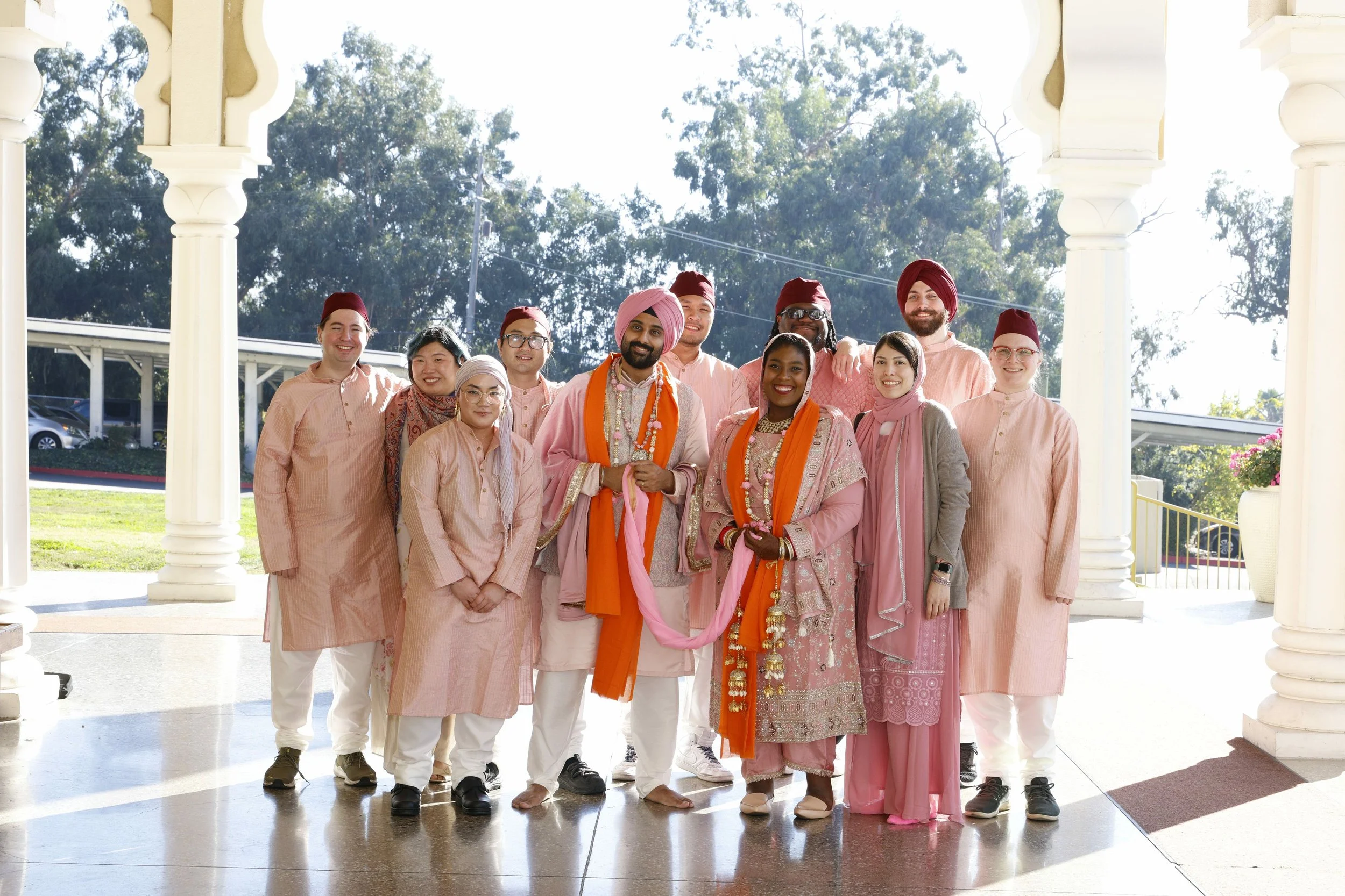 Group of men and women dressed in traditional Indian attire, standing outdoors under white columns with trees in the background, celebrating a cultural event.