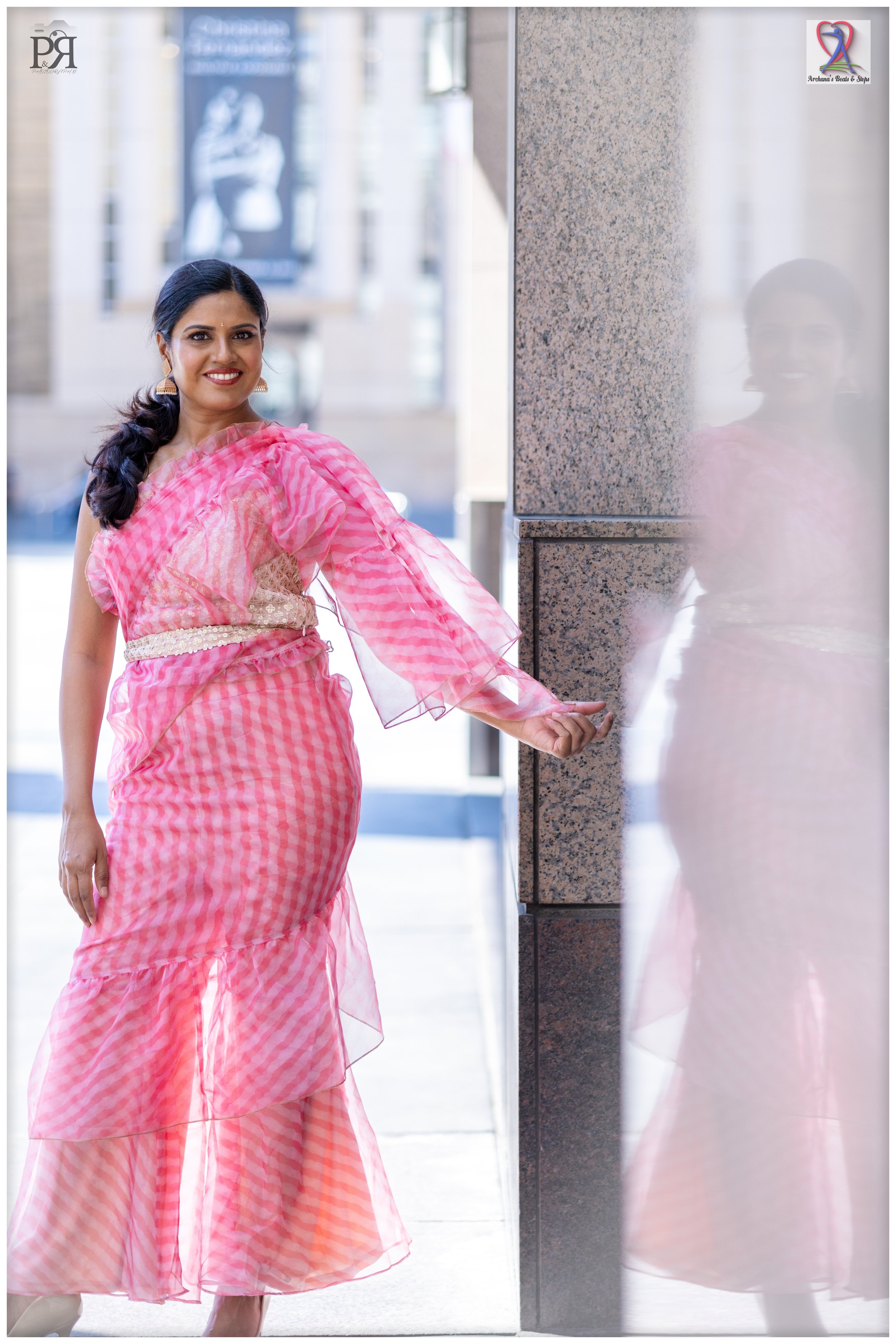 A woman dressed in a pink and white checkered saree, smiling and standing by a granite wall on a city street, with her reflection visible on a nearby glass surface.