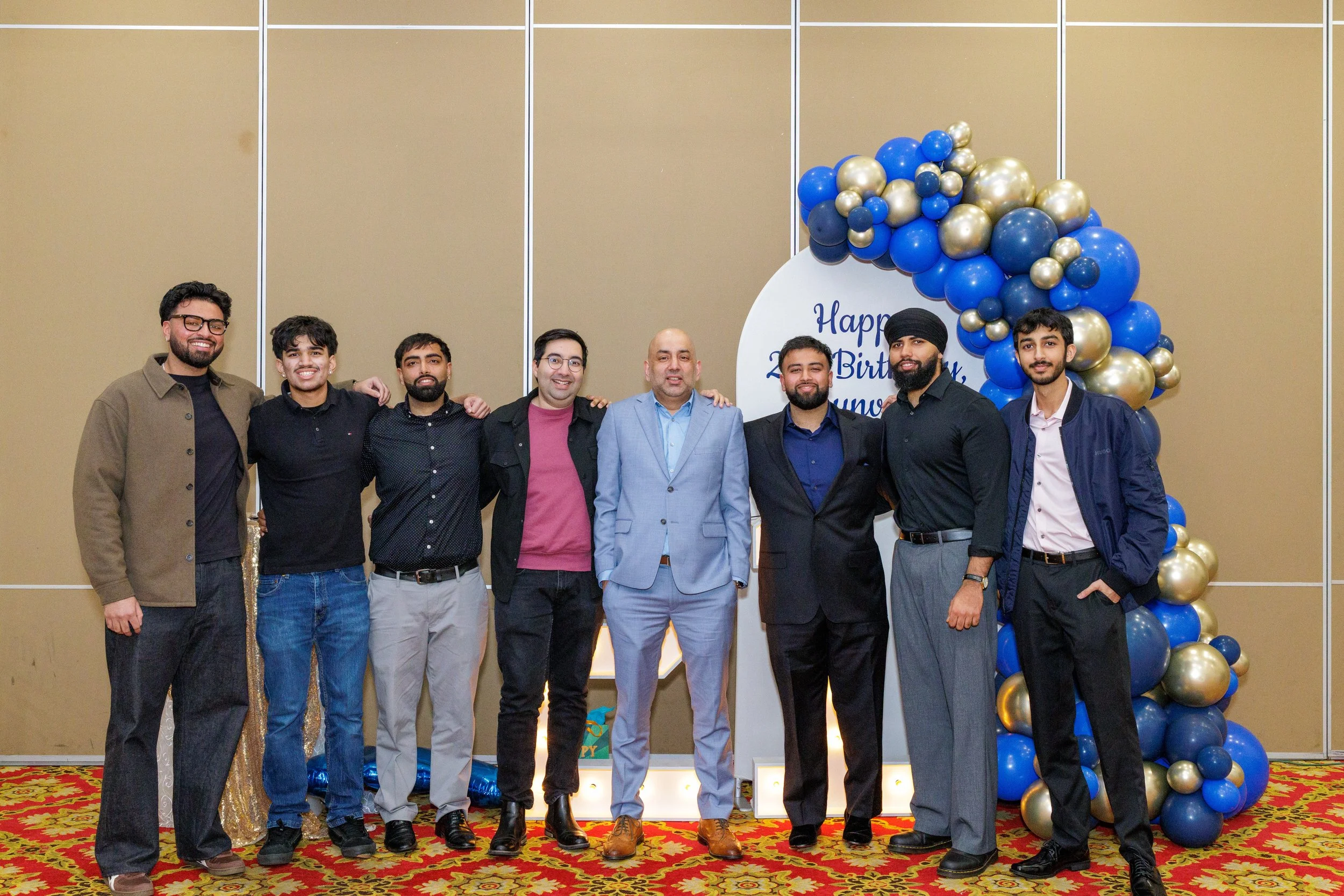 Group of nine men standing together at a birthday celebration. They are posing in front of a decorative balloon arch with blue, black, and gold balloons and a sign that says, 'Happy Birthday.' The men are dressed in casual and semi-formal attire and 