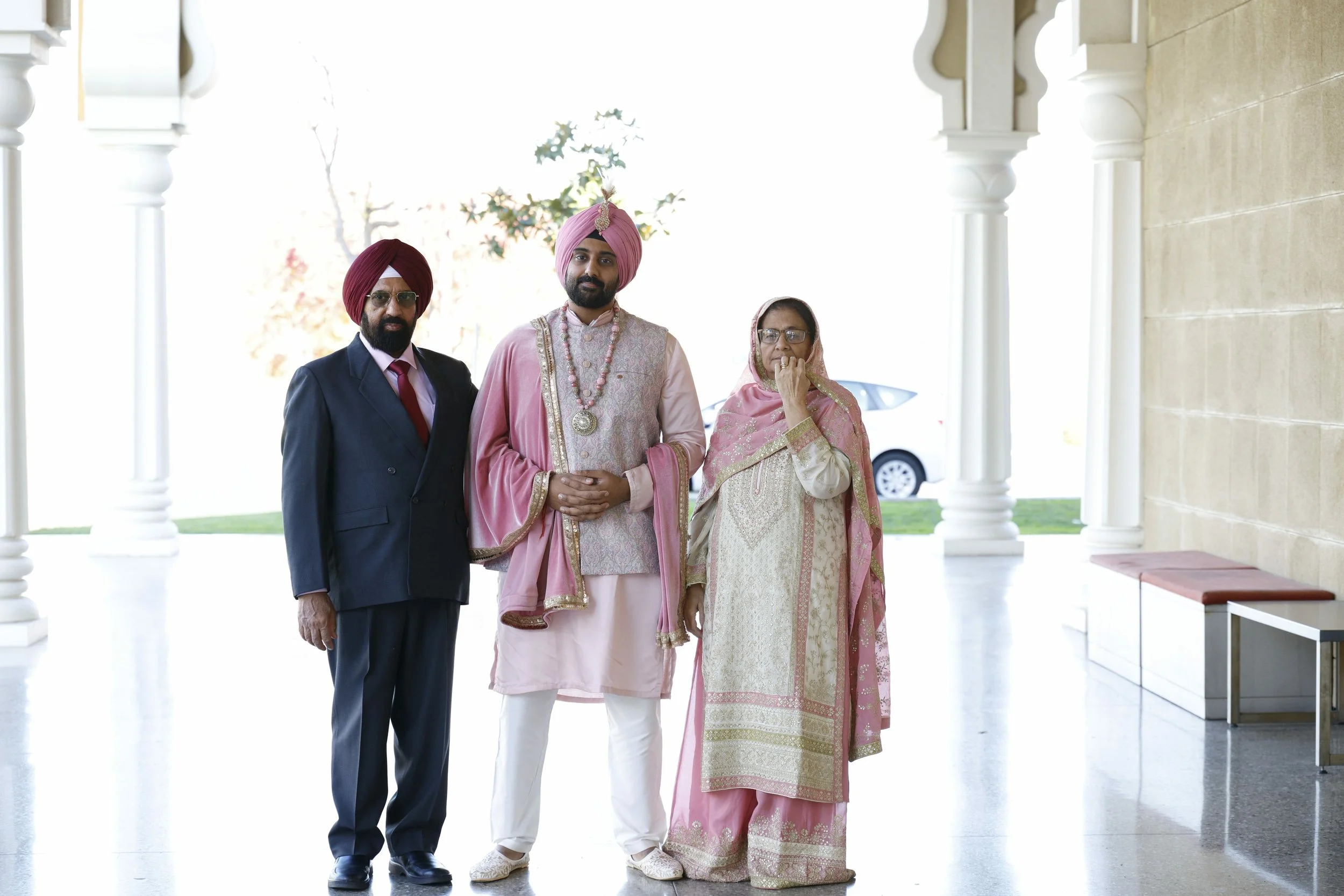 A group of four people at a wedding, including a groom wearing traditional Indian attire and his parents dressed in formal clothes, standing on a covered porch with white pillars and a car in the background.