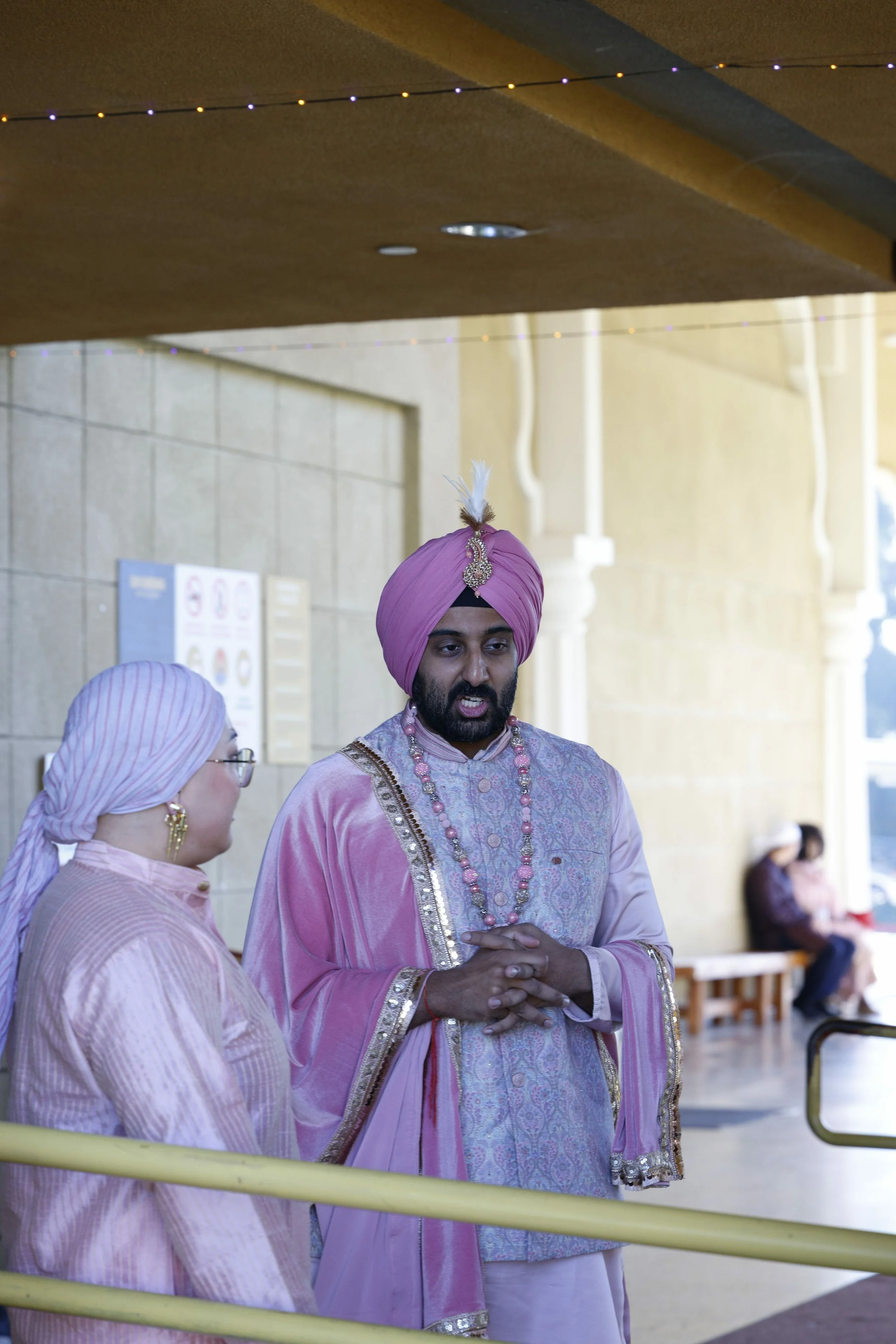 Man and woman dressed in traditional Indian attire, with the man wearing a pink turban and jewelry, standing and talking.