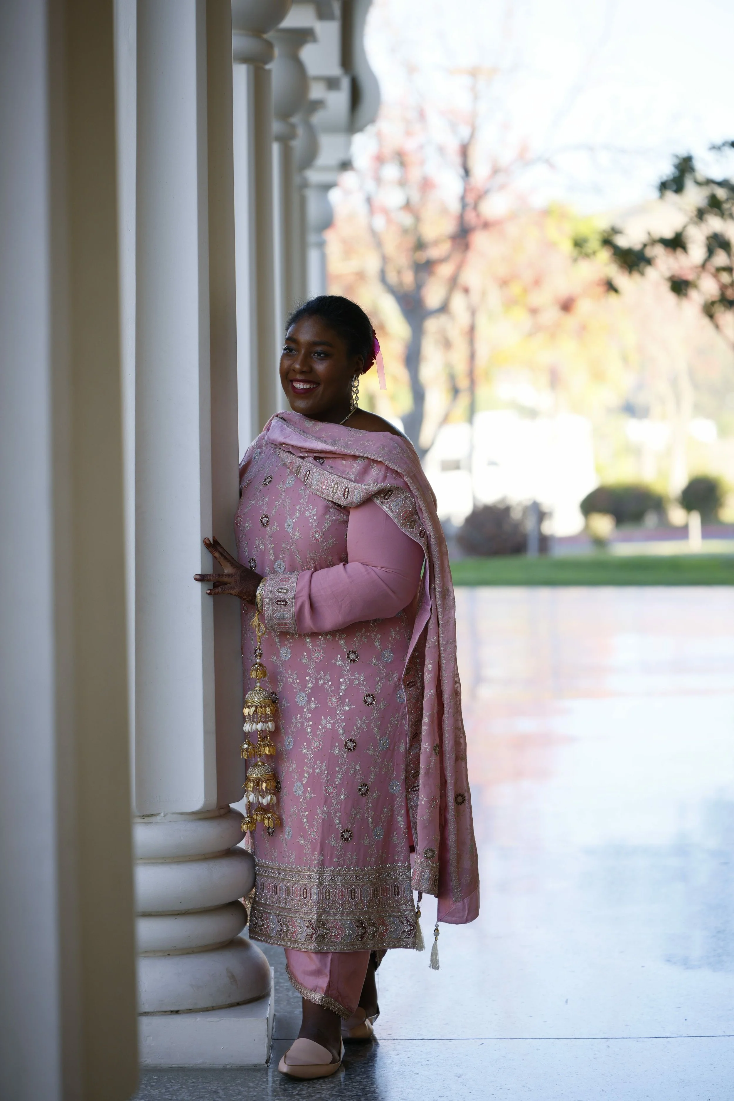 A woman in a pink traditional dress stands outside near white columns, smiling and looking to the side with trees and a lawn in the background.