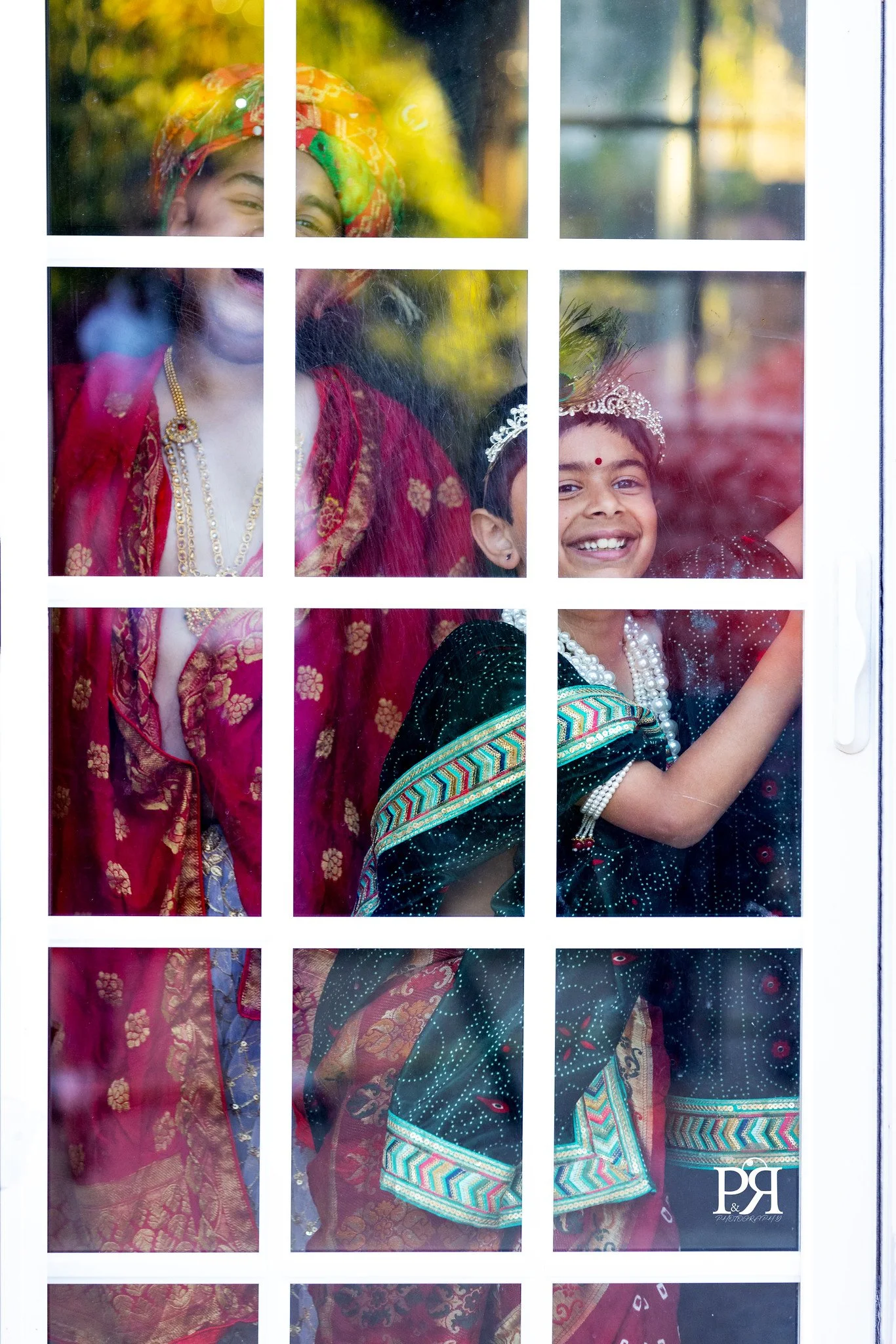 Smiling children dressed in colorful traditional Indian clothing, seen through a glass window with white grid pattern.