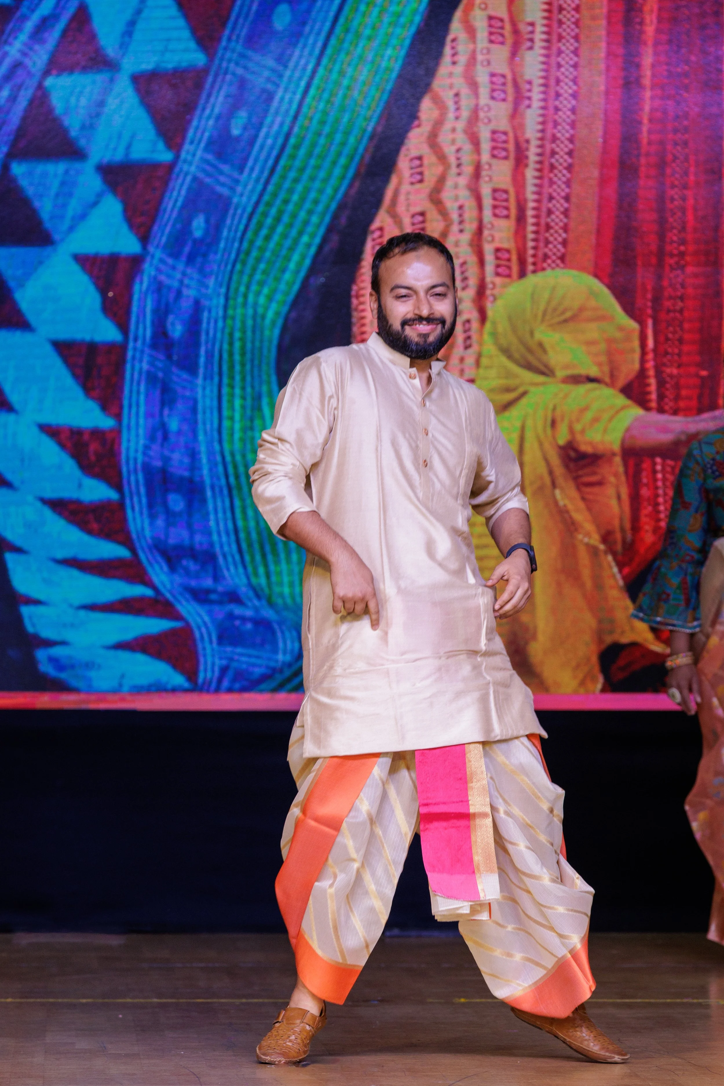 A man dancing on stage, wearing traditional Indian attire with a beige kurta and colorful dhoti, smiling, with a vibrant, colorful backdrop.