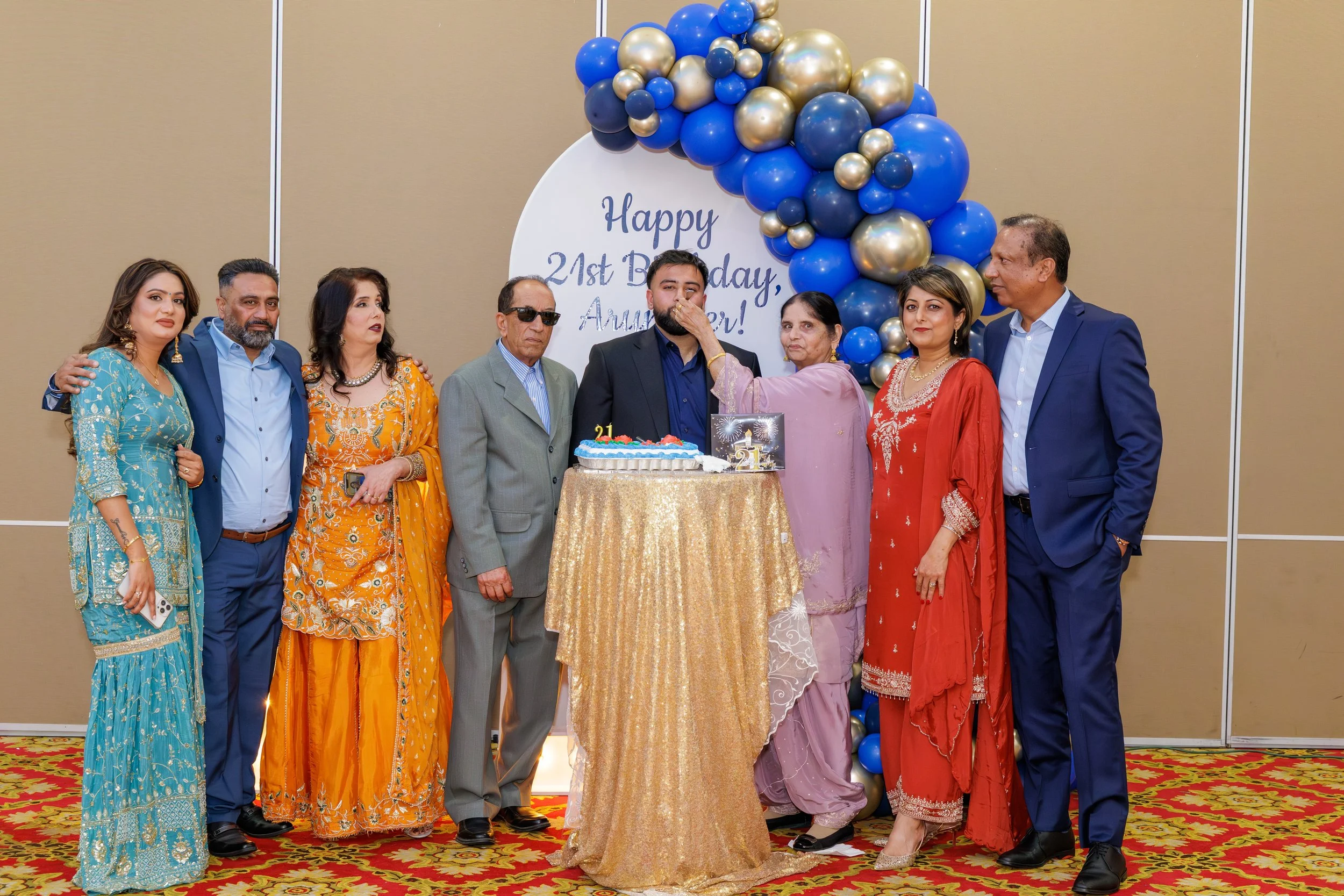 A group of nine people dressed in colorful traditional and formal attire celebrating a birthday with a decorated backdrop, balloons, and a cake in the middle. The backdrop says 'Happy 21st Birthday, Anum Ver!'