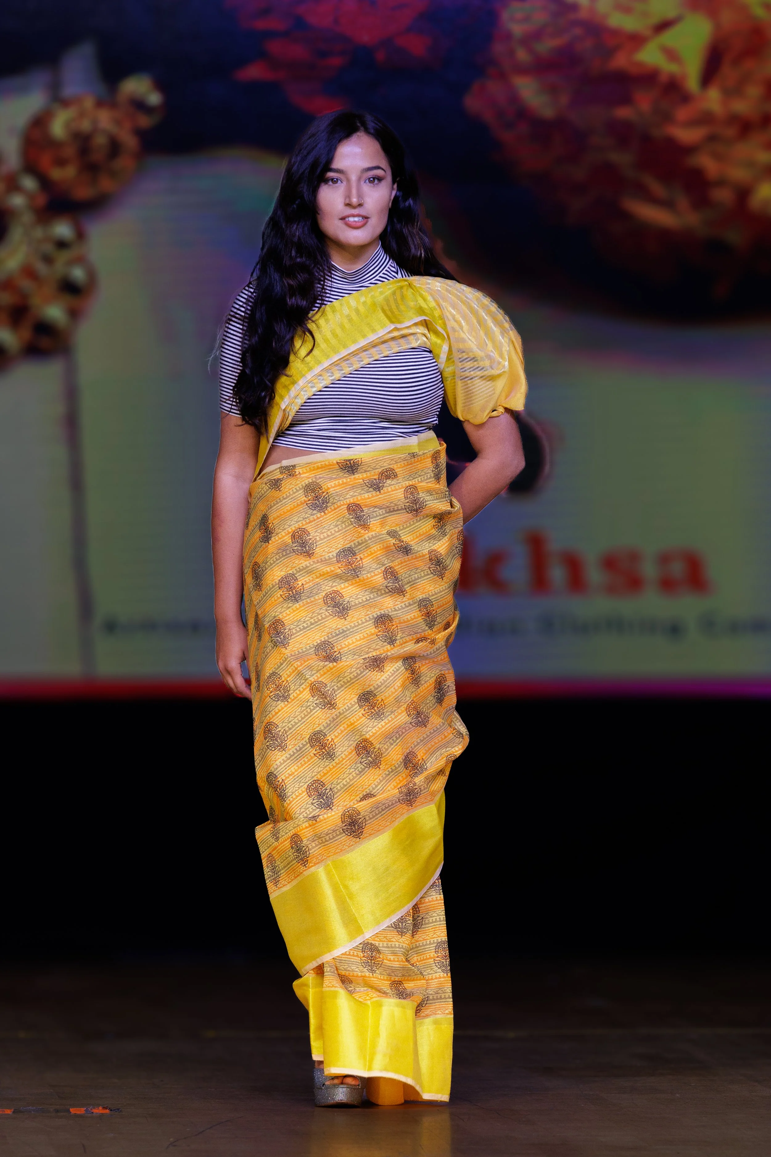 A woman walking on a runway wearing a traditional yellow saree with black and white striped blouse, with long dark hair.