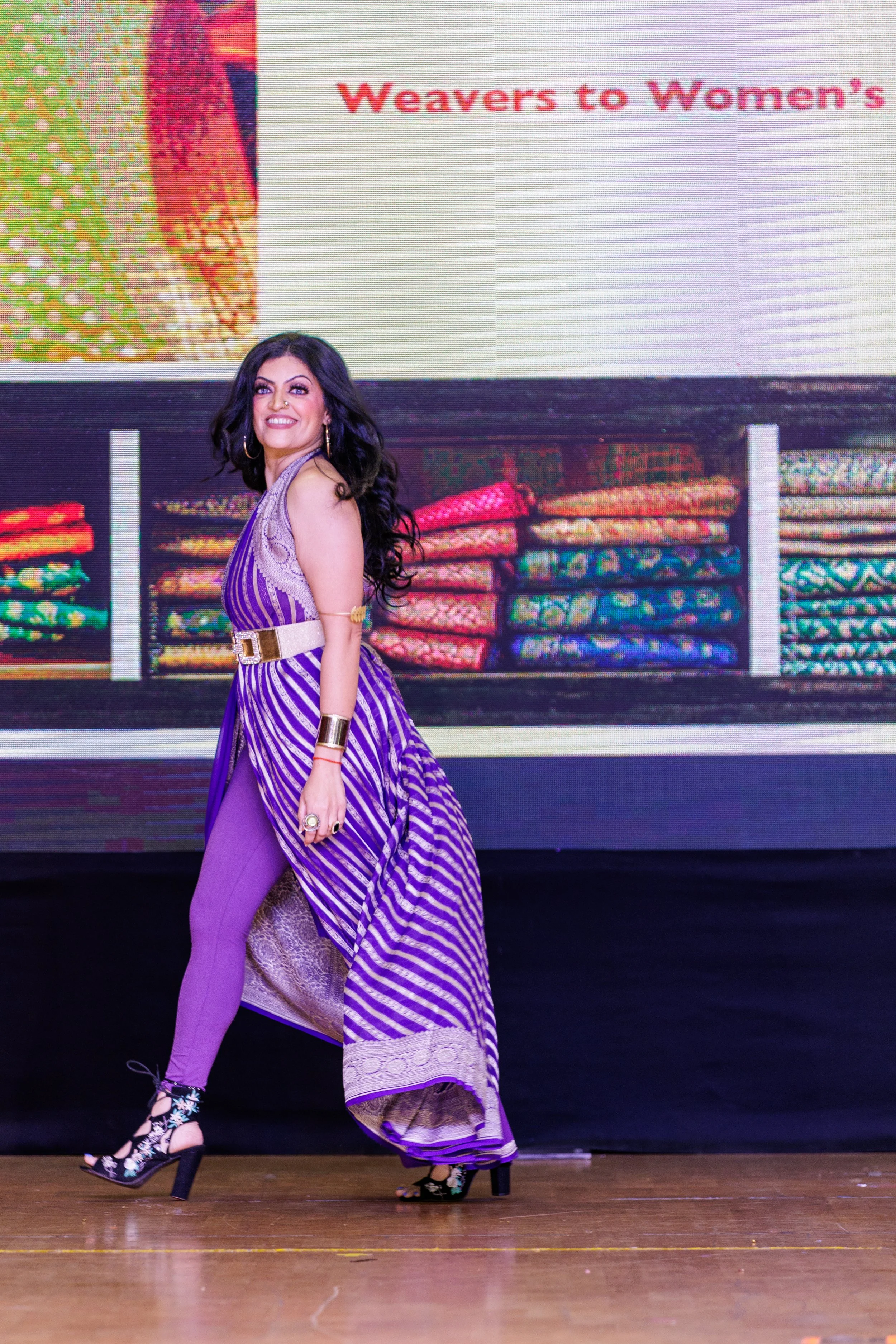 A woman walking on stage wearing a purple and white striped dress with high heels, and colorful accessories, in front of a digital screen displaying text 'Weavers to Women's' and images of folded fabrics.
