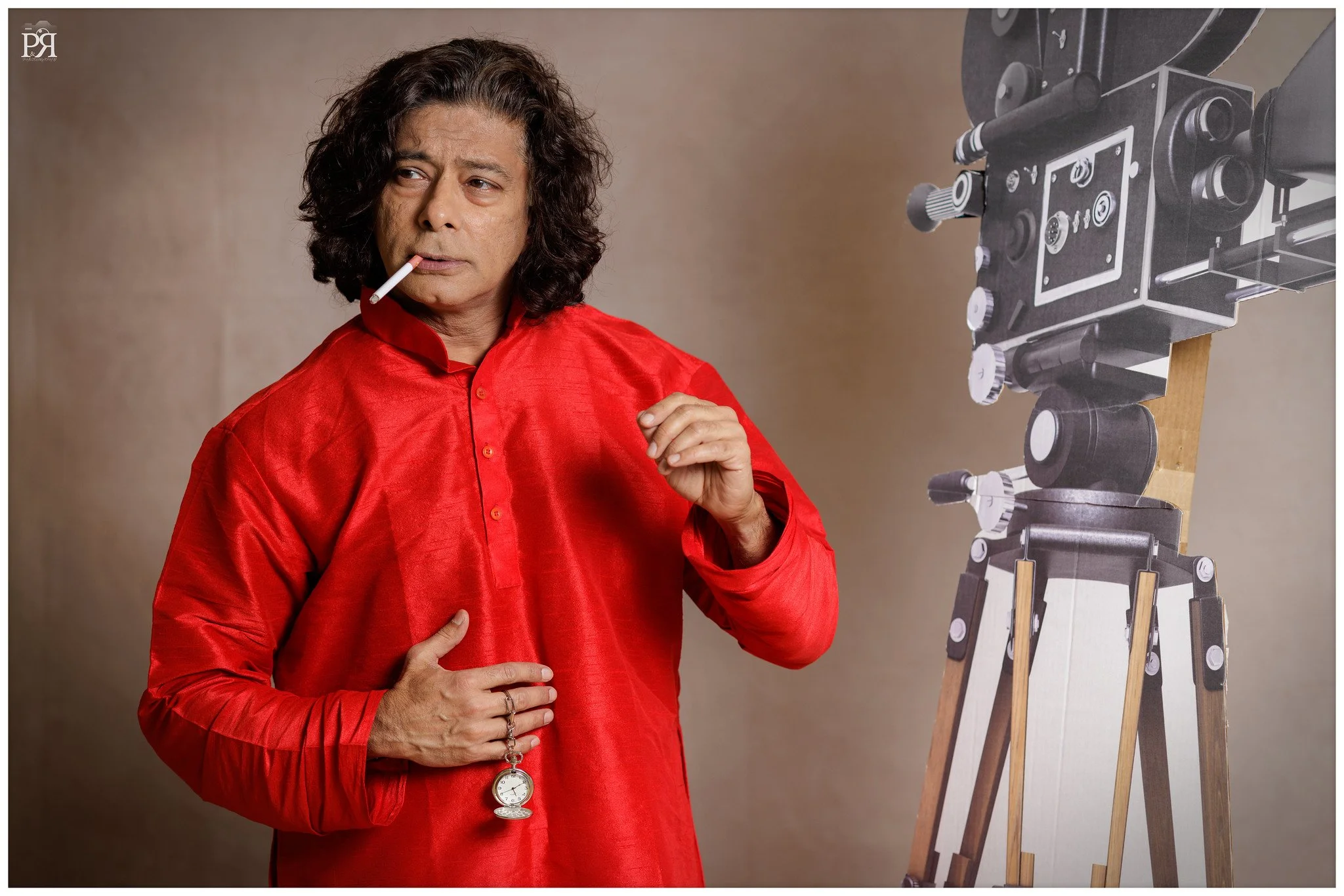 A man wearing a red shirt holding a pocket watch and a cigarette, standing next to a large vintage camera mounted on a tripod.
