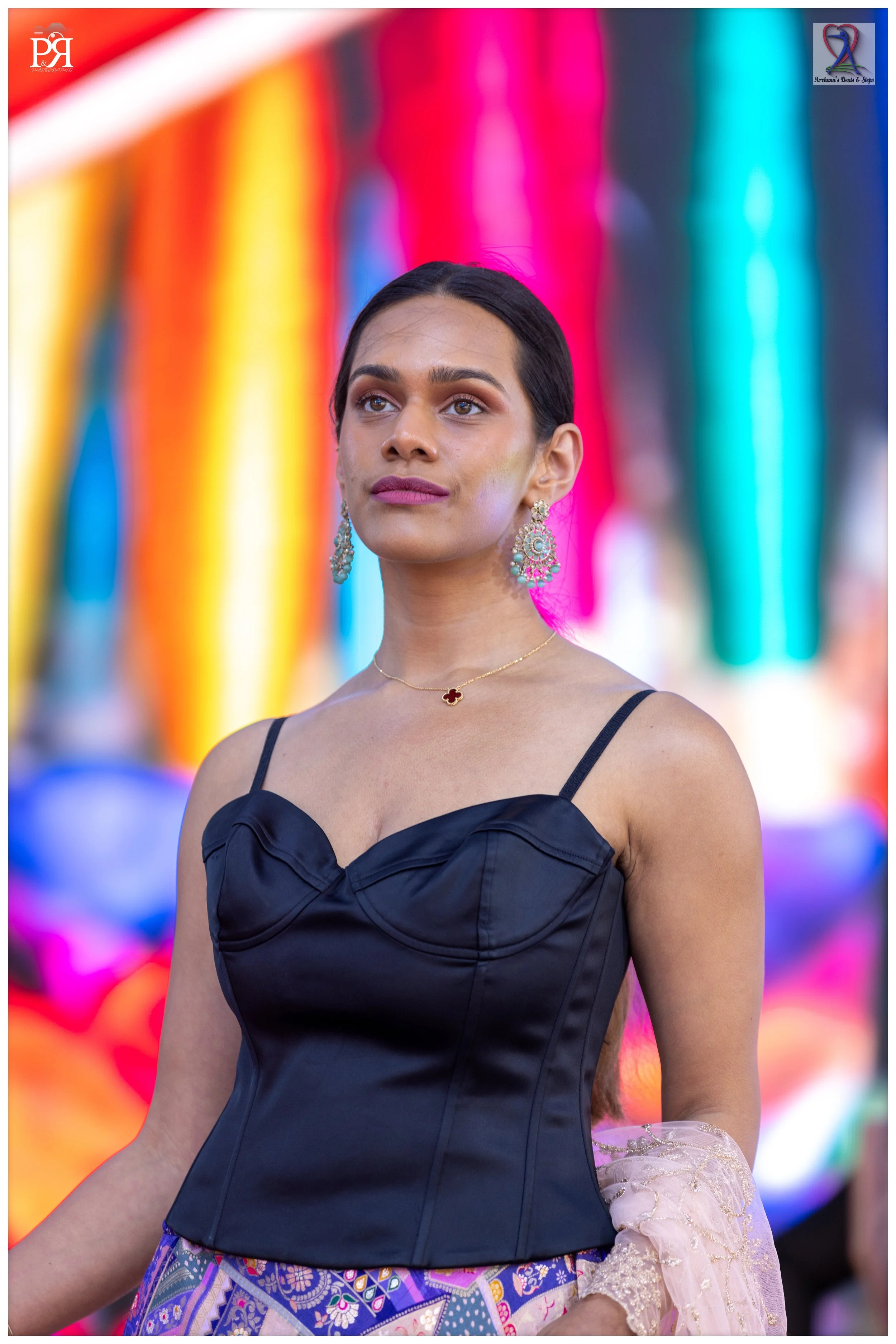 A woman with dark hair and medium skin tone wearing a black satin top, large earrings, a gold necklace, and holding a pink fabric, standing in front of a colorful blurred background.