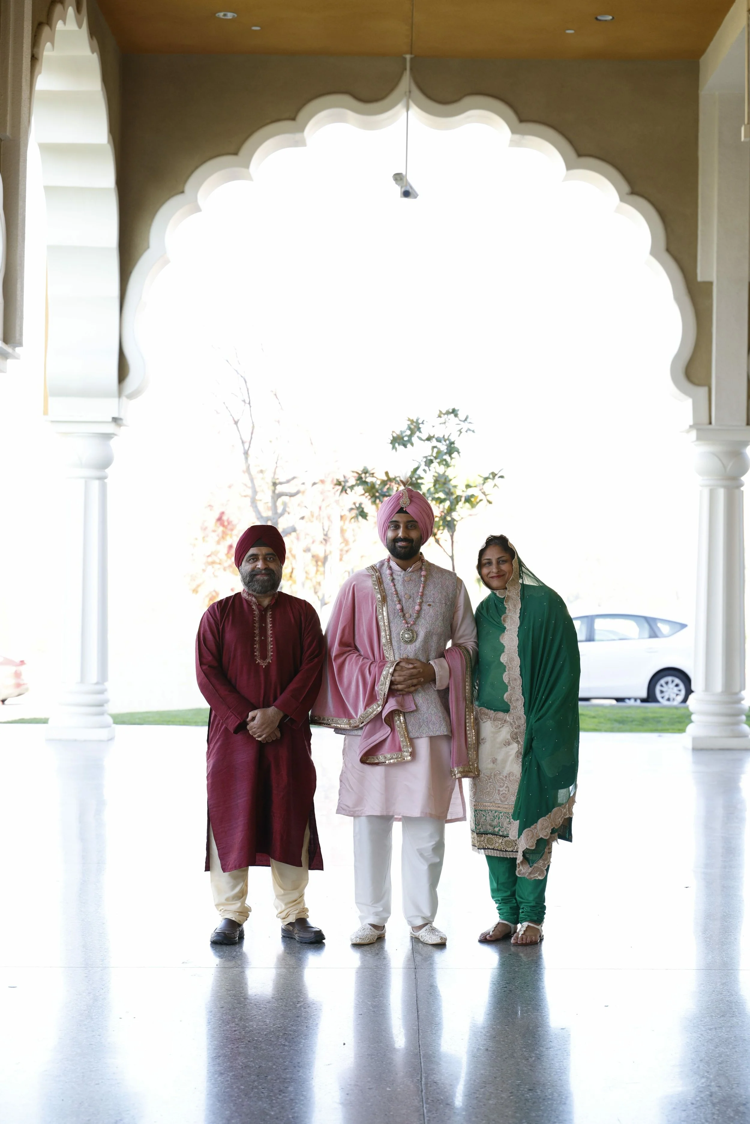 Three people in traditional Indian attire standing under an ornate archway, with a car and trees in the background.