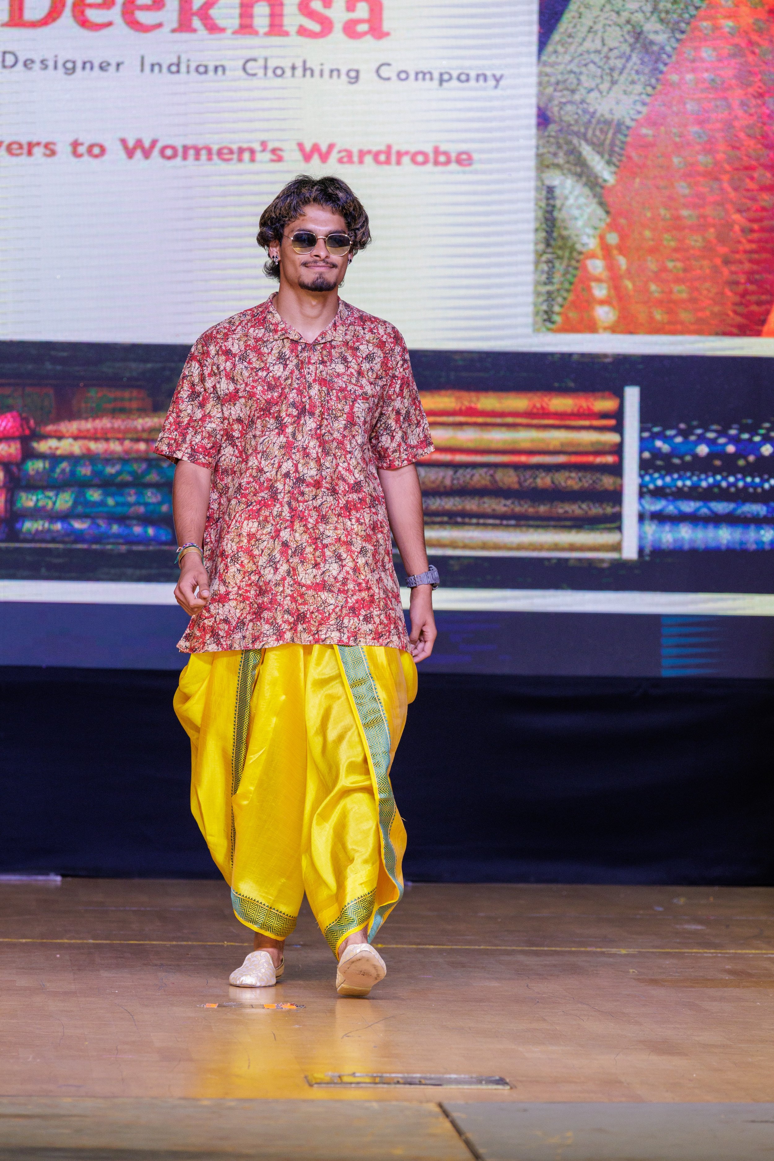 A man walking on a stage wearing a traditional Indian outfit with a red and gold patterned kurta and bright yellow dhoti pants, with books or fabrics displayed behind him on a screen.
