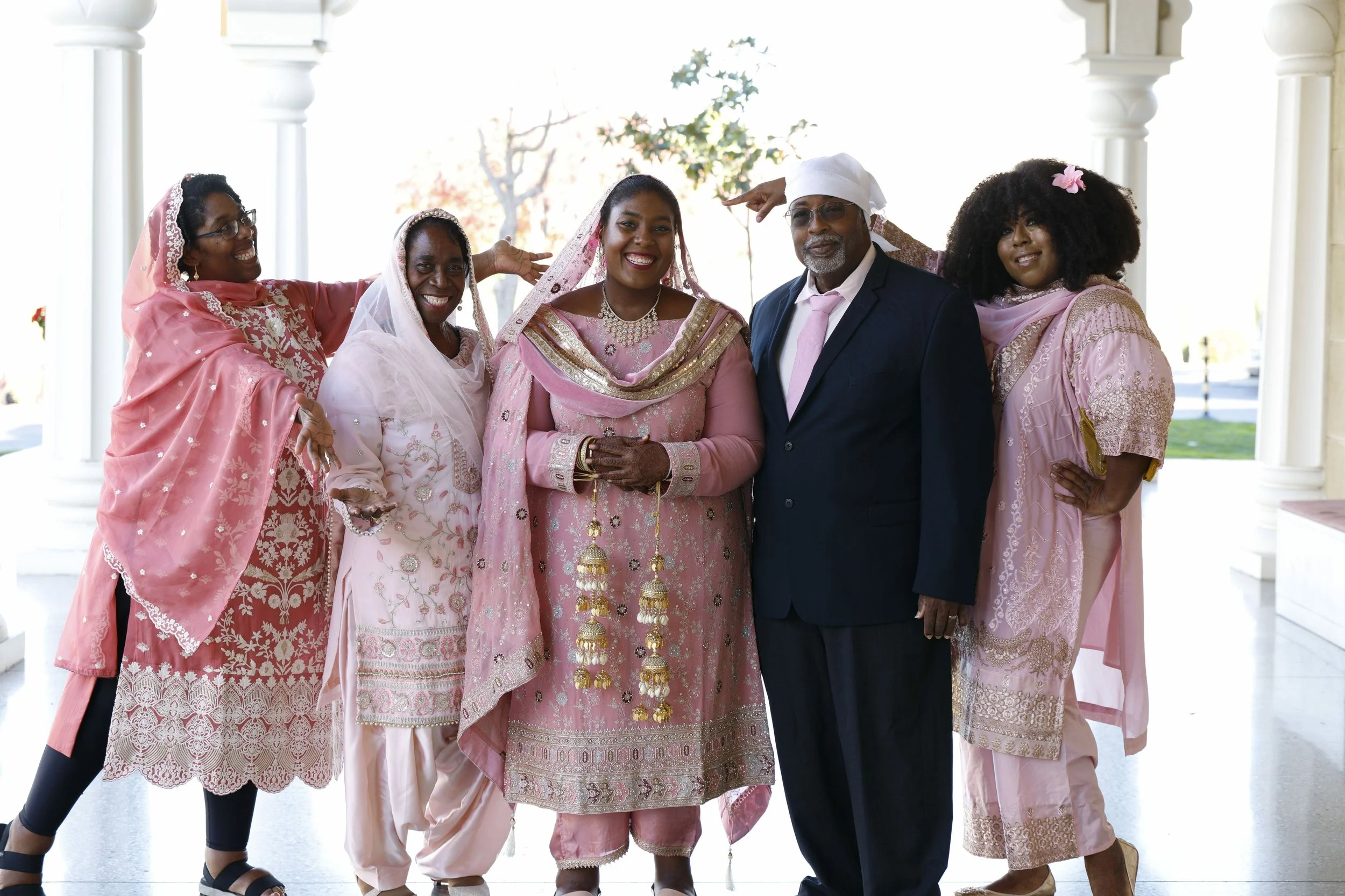 A group of six people dressed in traditional South Asian attire, standing together in a bright, outdoor setting. They are smiling and appear to be celebrating a special occasion.
