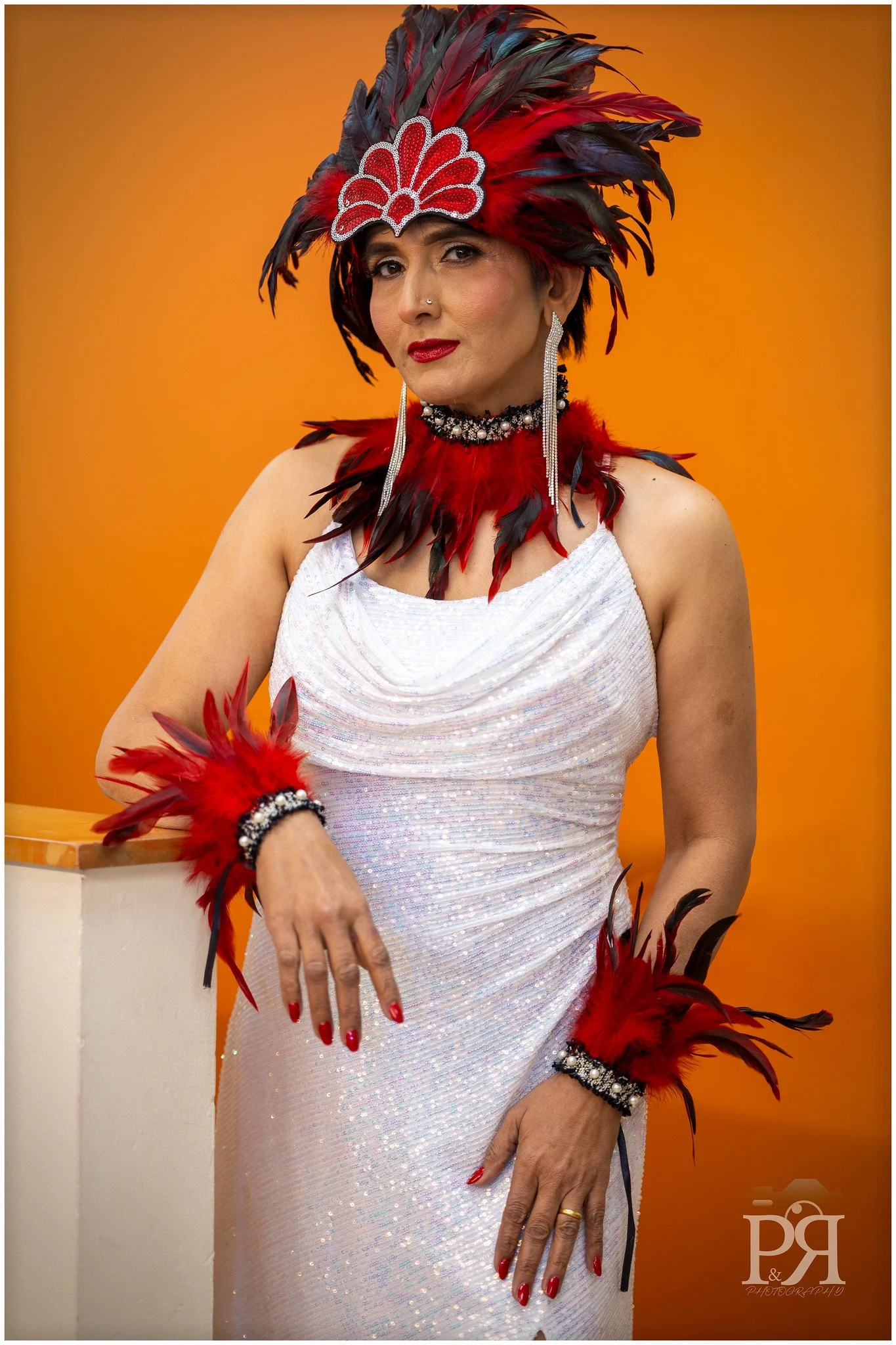 A woman dressed in a sparkly white gown with red feather accessories, wearing a large feathered headdress, jewelry, and bright red lipstick, posing against an orange background.
