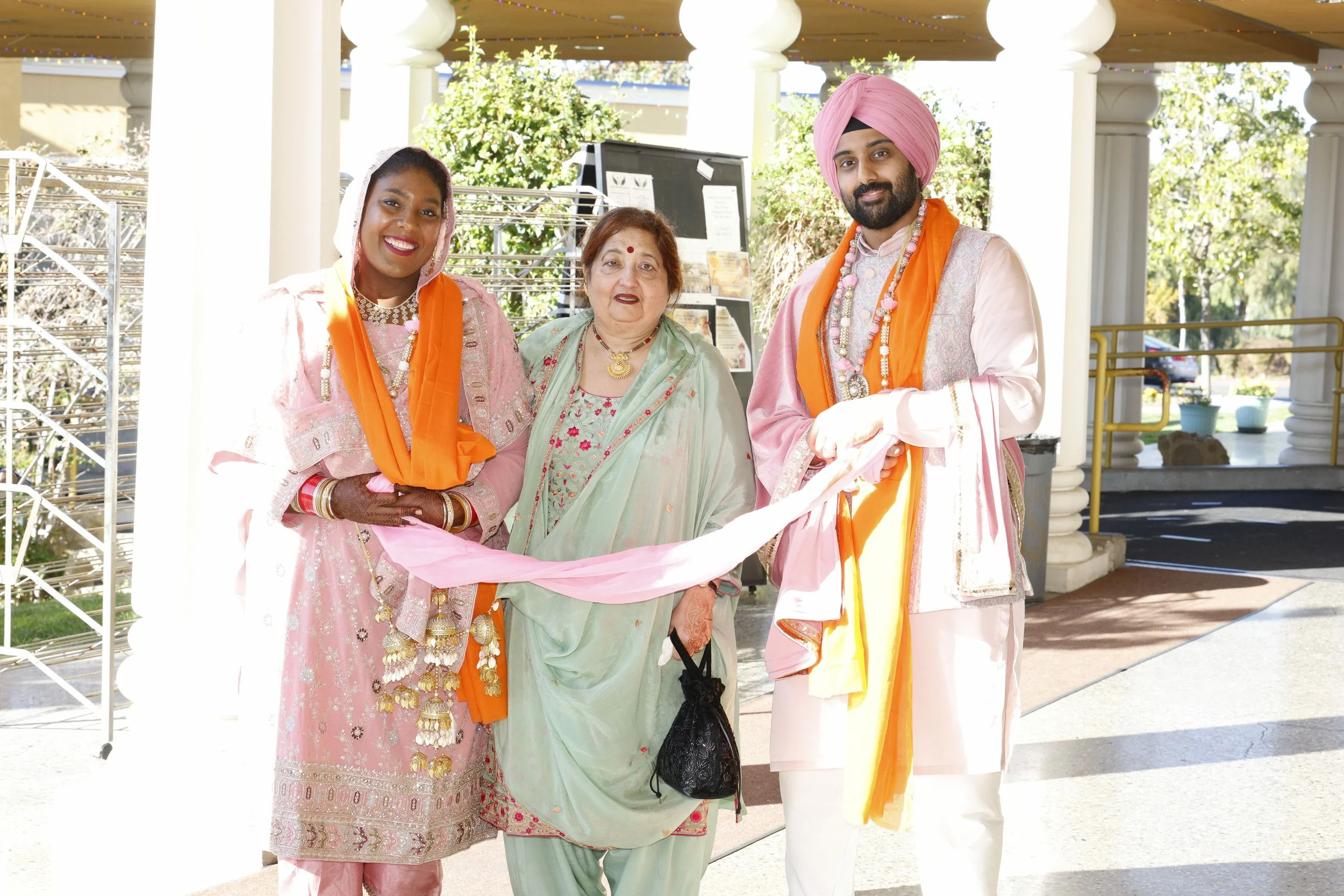 Four people dressed in traditional Indian attire participate in a cultural celebration outdoors, holding a pink cloth together, with buildings and trees in the background.