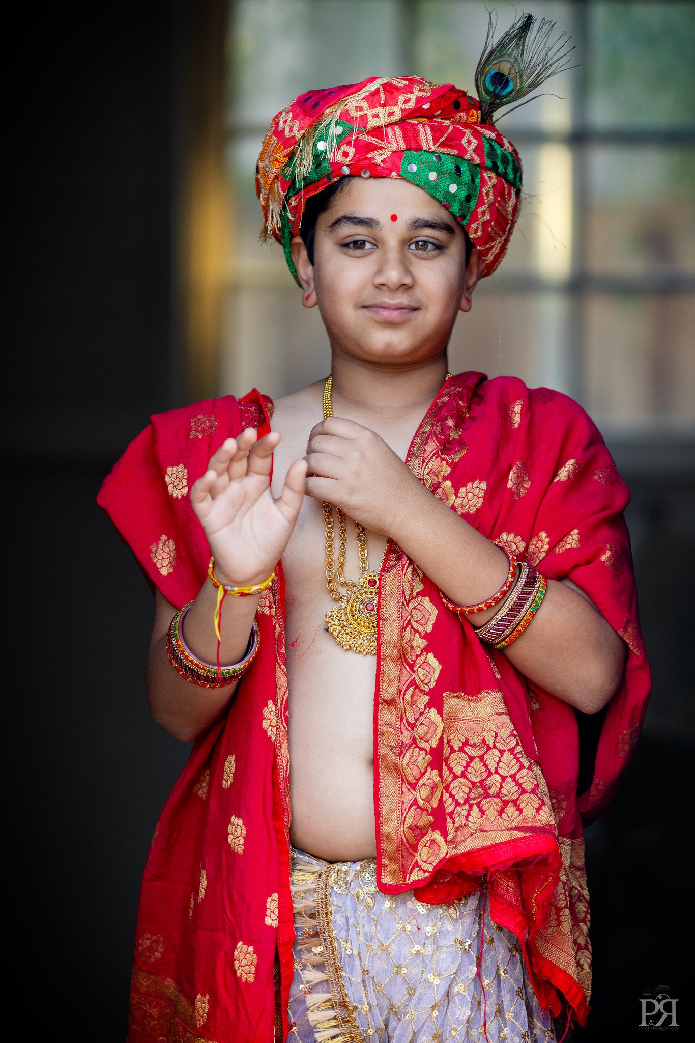 A young boy dressed in traditional Indian attire, with a red and gold embroidered turban, red shawl, and jewelry, standing against a blurred background.