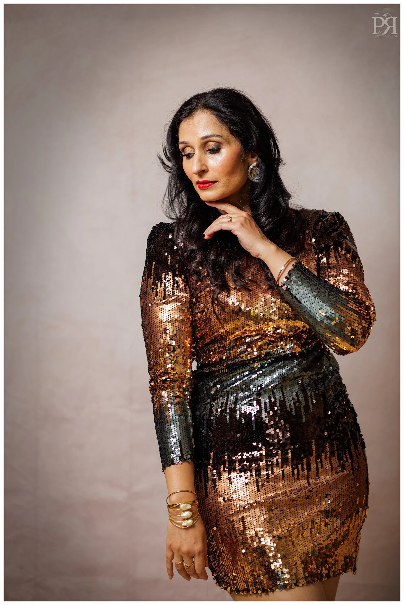 A woman in a long sleeve, sequined, gold and black dress with earrings and bracelets, posing with her hand near her chin, looking down.