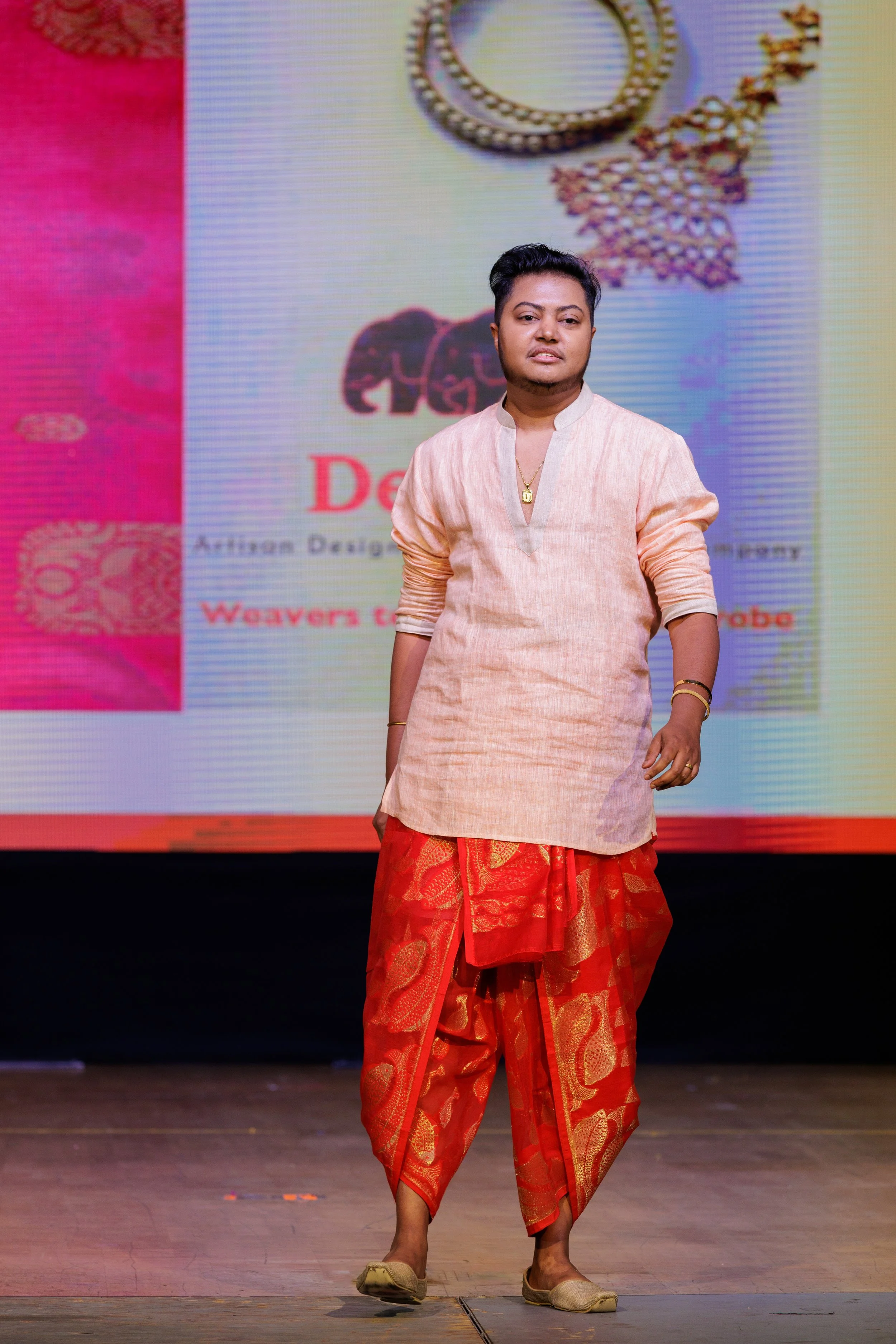 A person walking on stage in front of a large screen displaying jewelry and fashion design advertisements. The person is dressed in traditional Indian attire, including a peach kurta and red saree with gold patterns, and is wearing simple jewelry.