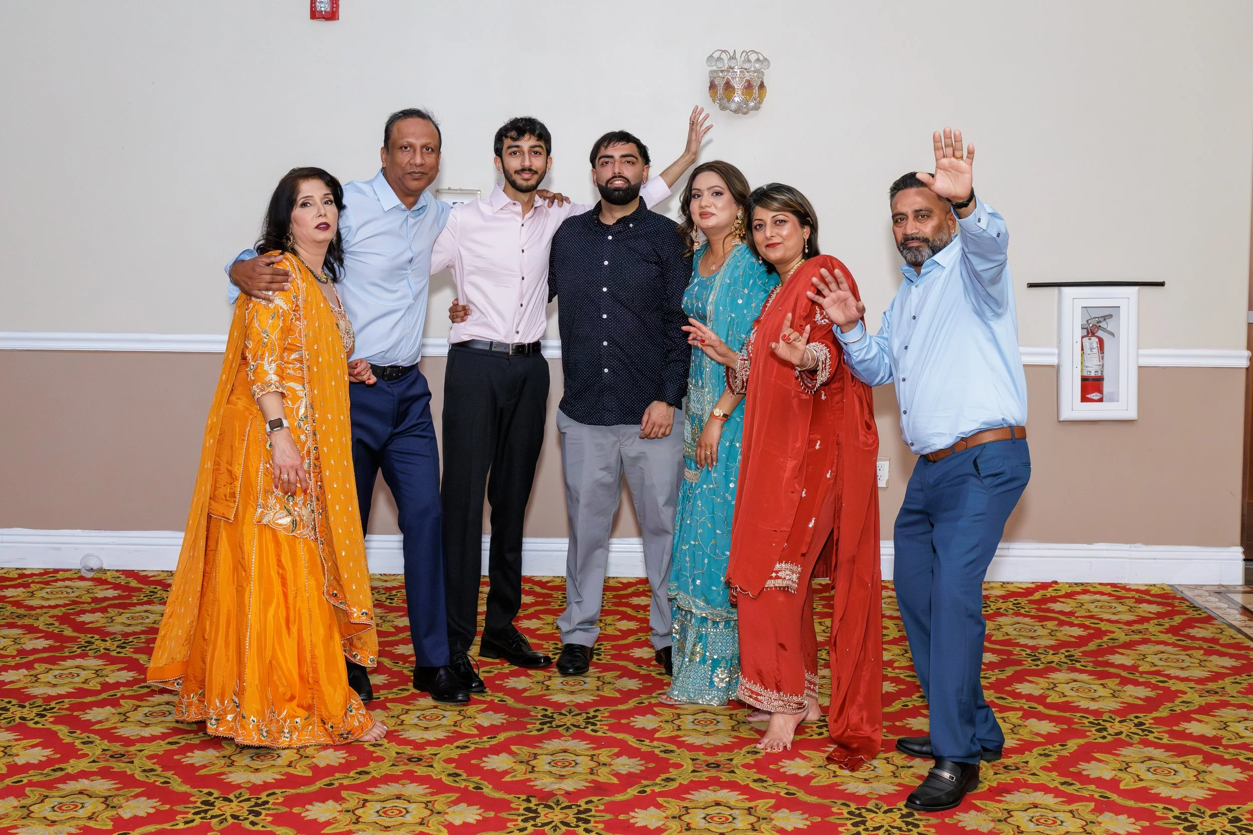 Group of eight people in colorful traditional and formal attire posing indoors on a decorative carpet. They are smiling, some waving, and standing close together against a beige wall.