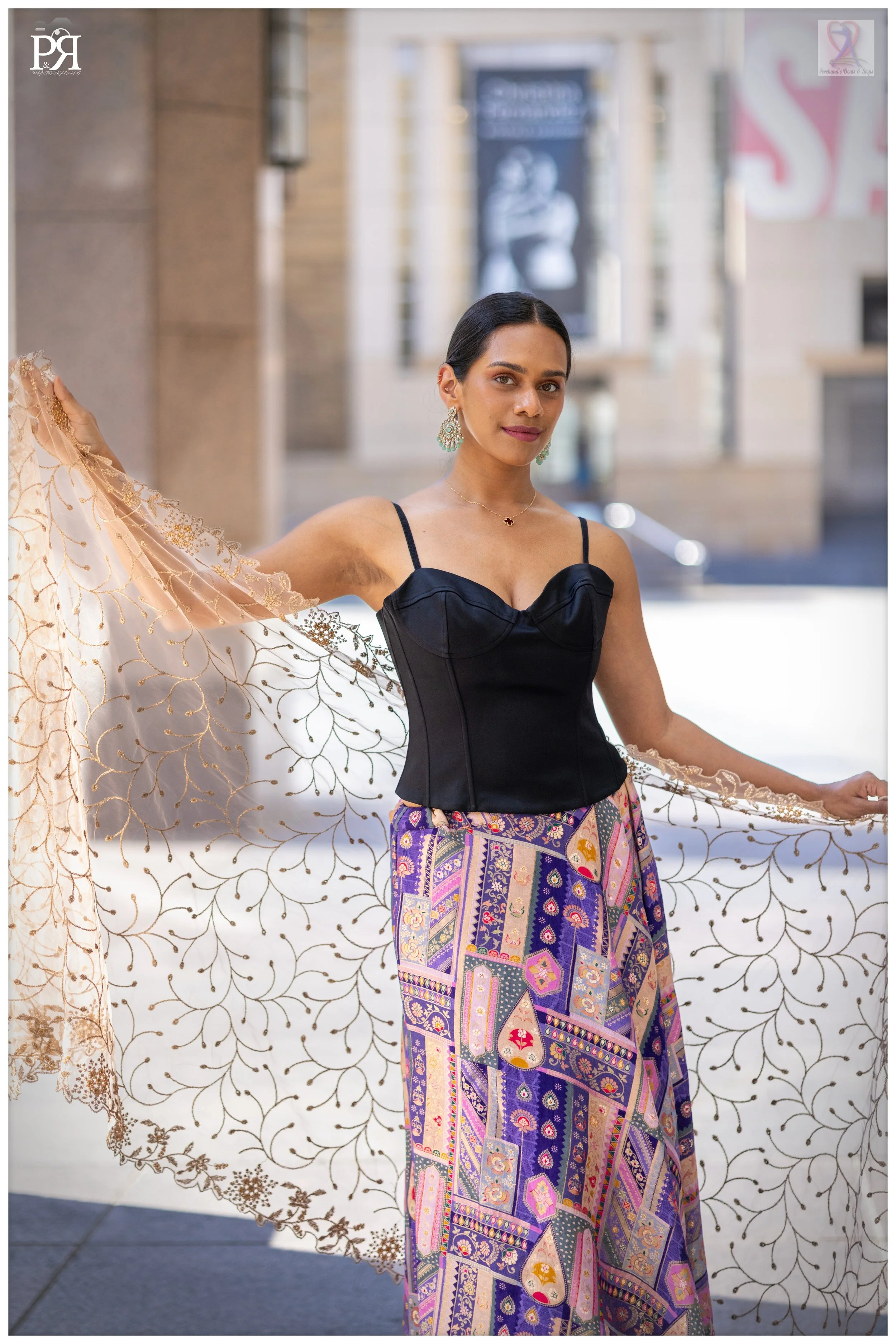 A woman with dark hair styled back, wearing earrings, a black sleeveless top, and a colorful patterned skirt, holding a sheer embroidered shawl outdoors in an urban setting.