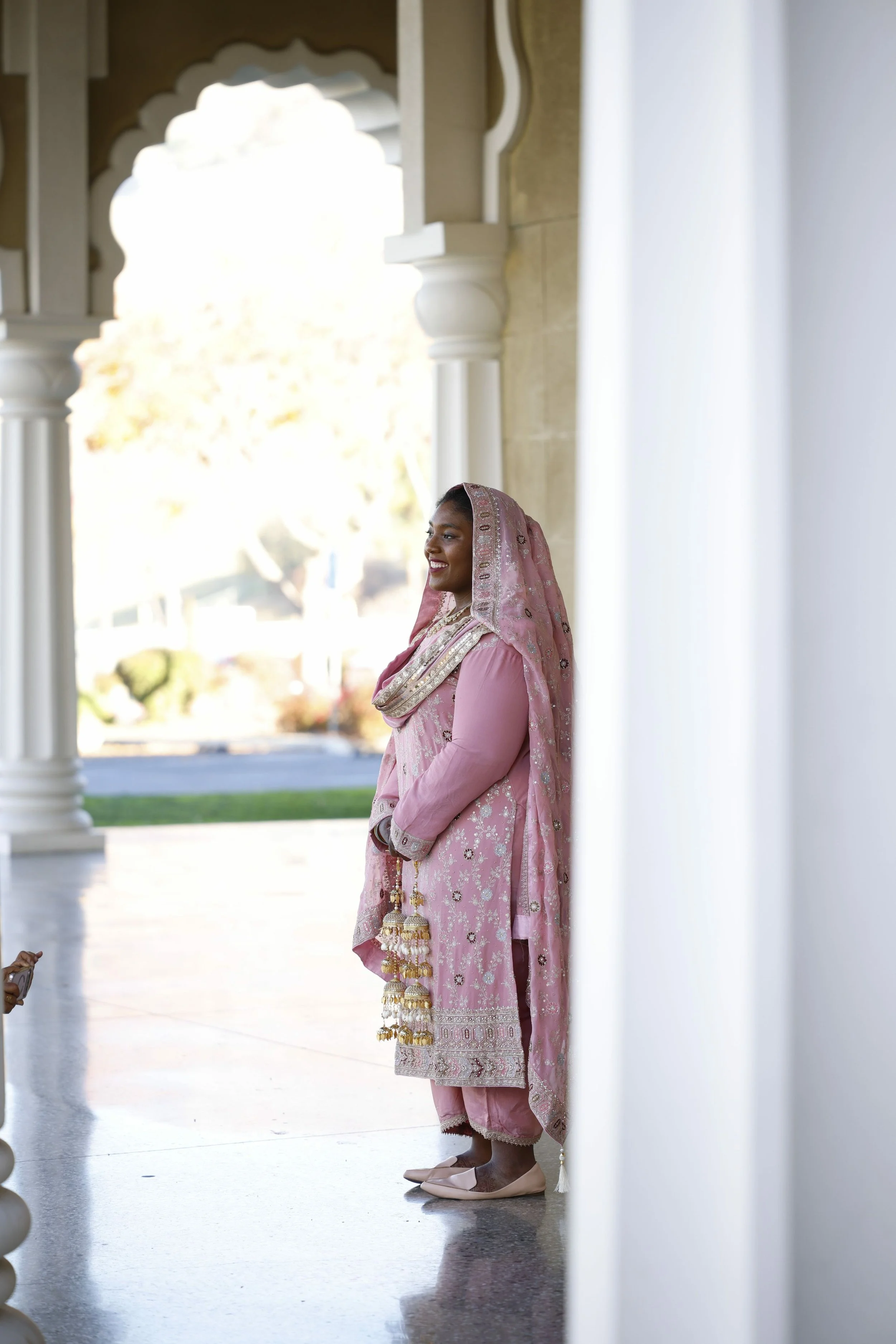 Smiling woman in traditional pink Indian attire standing in a corridor with white columns and arches.