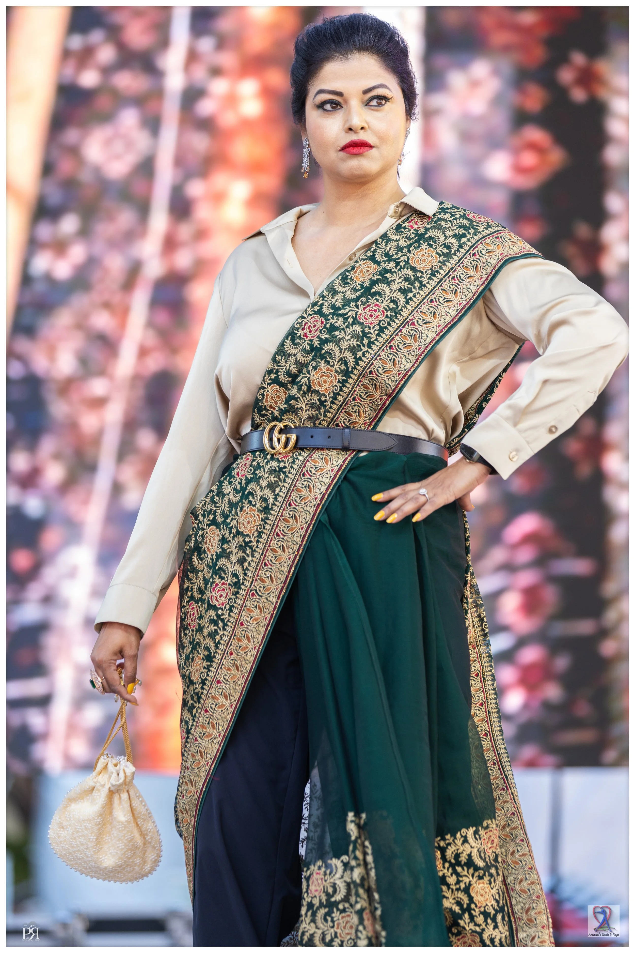 Woman wearing a traditional saree with intricate embroidery, a beige shirt, and a Gucci belt, holding a small pouch in her left hand and standing confidently.
