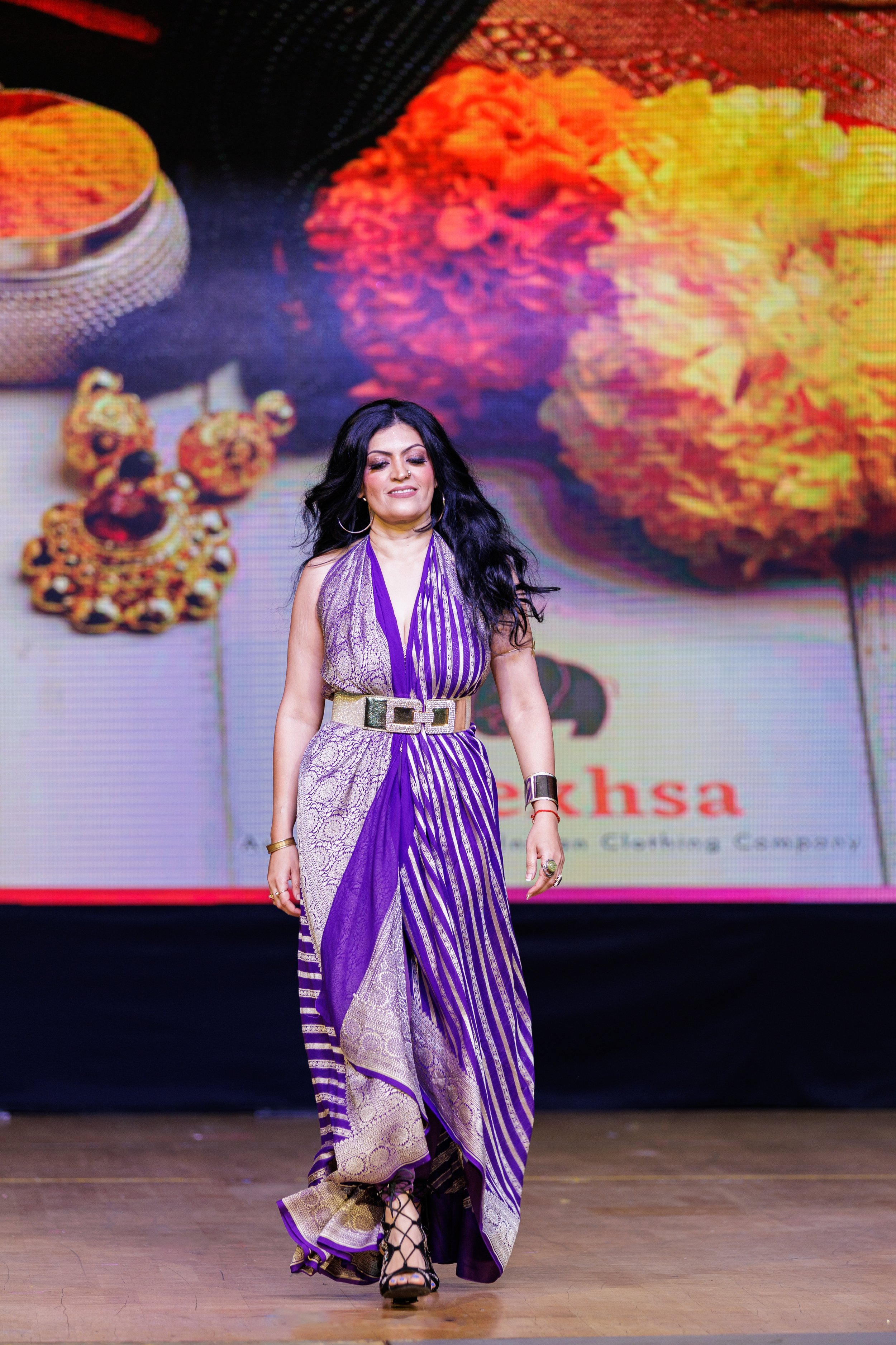 Woman in a purple and gold saree walking on stage with a colorful background of jewelry and flowers.