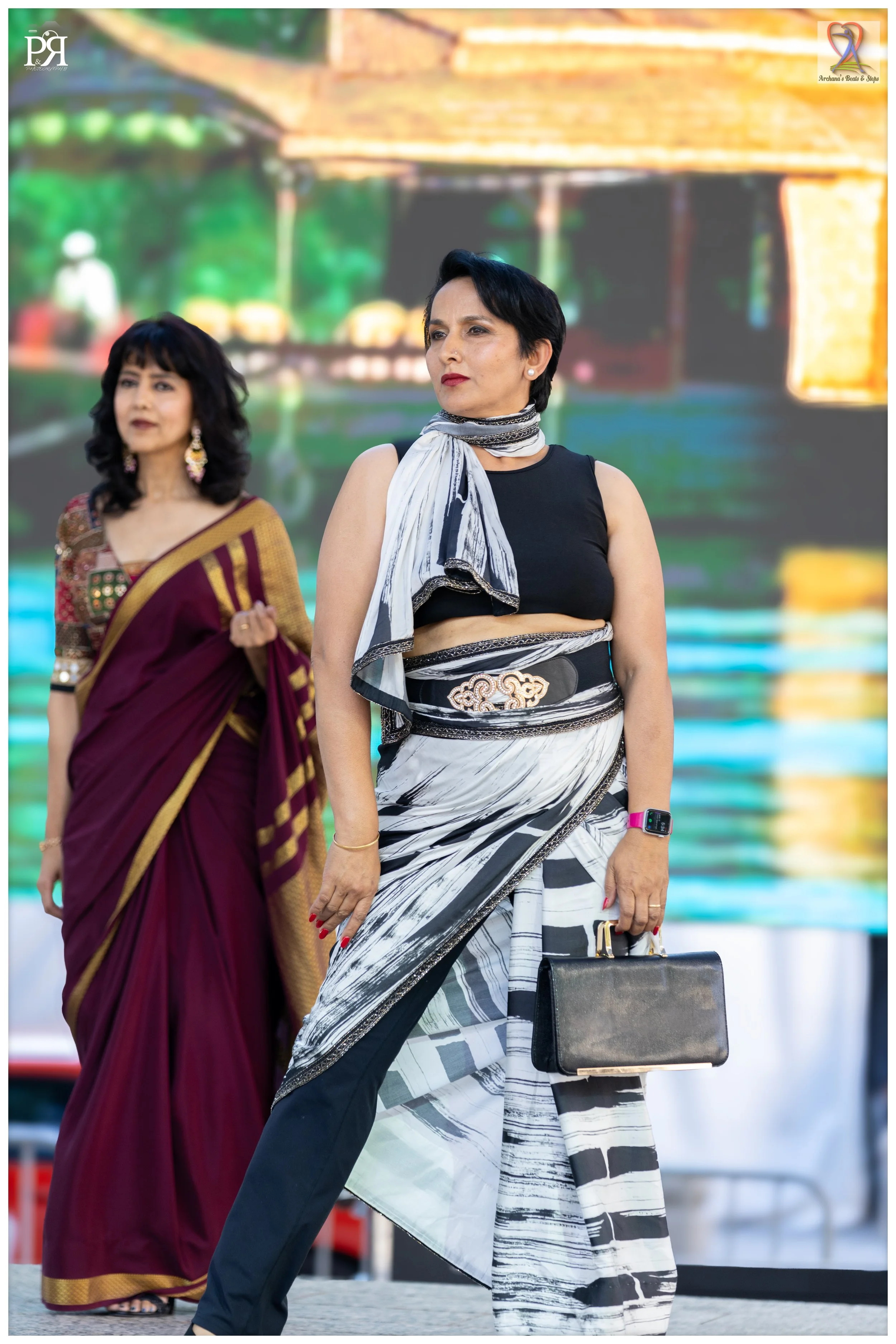 Two women standing on stage, one in a traditional saree and the other in a fashionable black and white ensemble, holding a black handbag, with a blurred colorful background.
