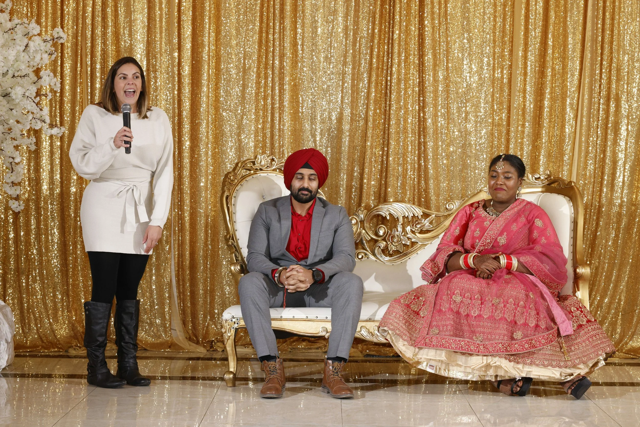 A woman in a white sweater dress and black boots standing with a microphone, beside a man in a gray suit with a red turban, and a woman in a pink traditional saree seated on a decorated set with gold curtains.