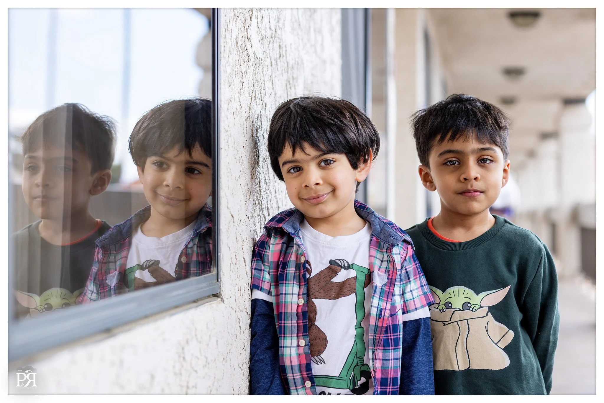 Three young boys standing outside near a textured wall and window, with their reflections visible in the glass.