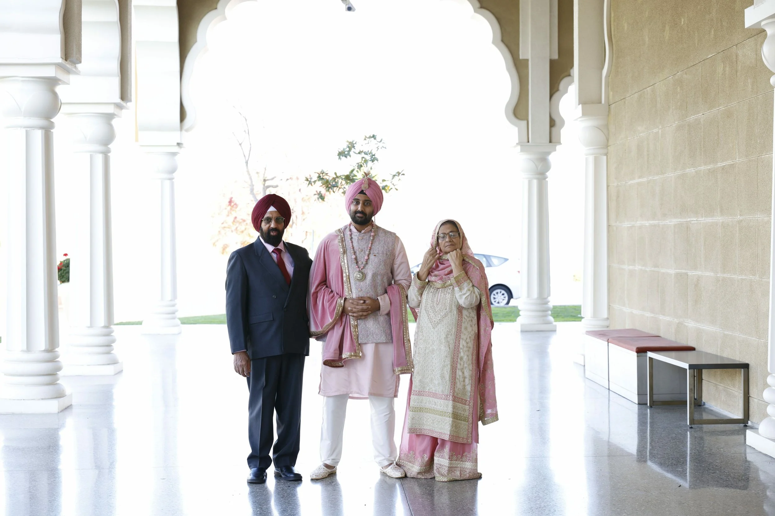 A family of three, dressed in traditional Indian wedding attire, standing under a white architectural archway with detailed columns on a bright day.