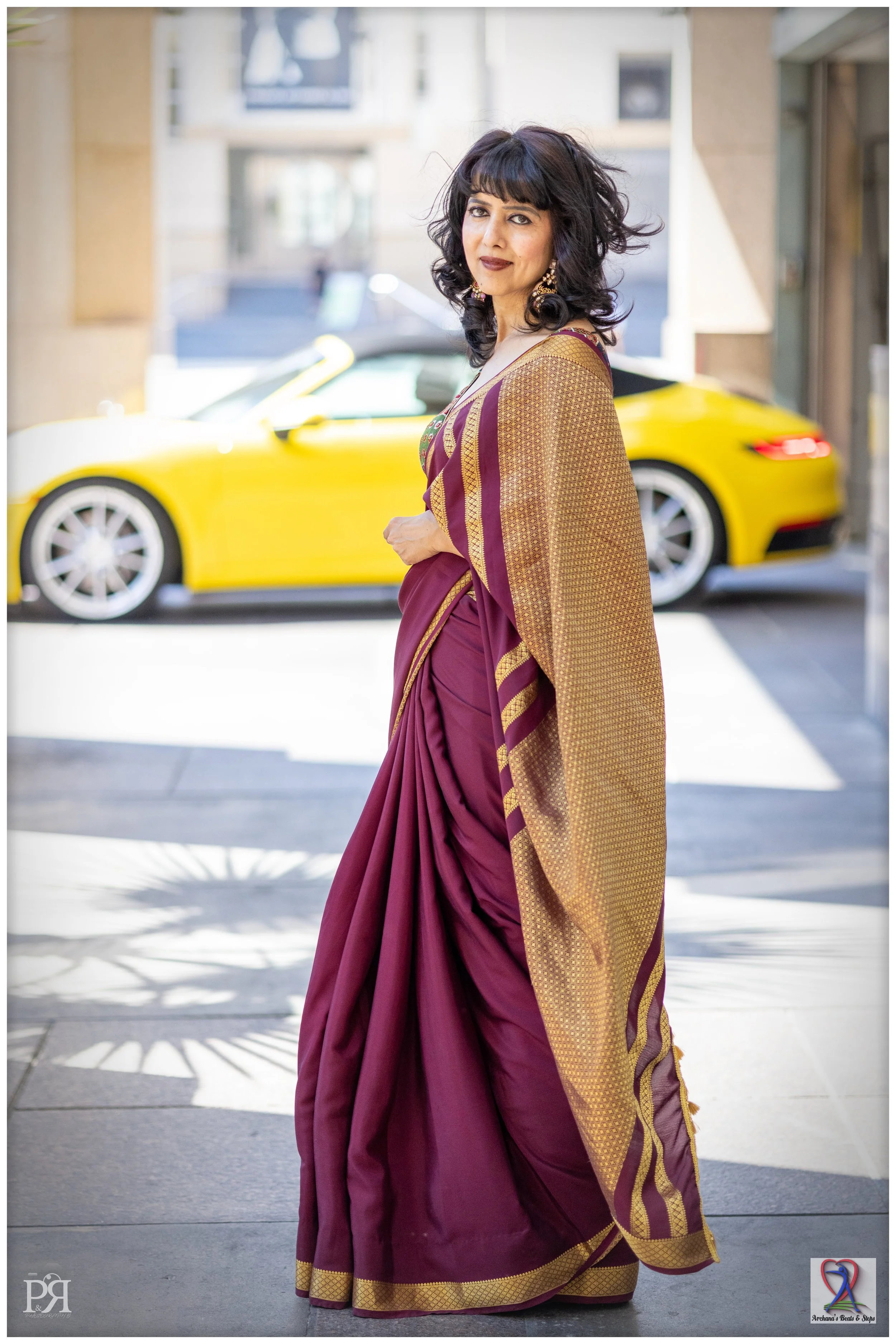 A woman in a purple and gold saree standing on a sidewalk with a yellow sports car in the background.