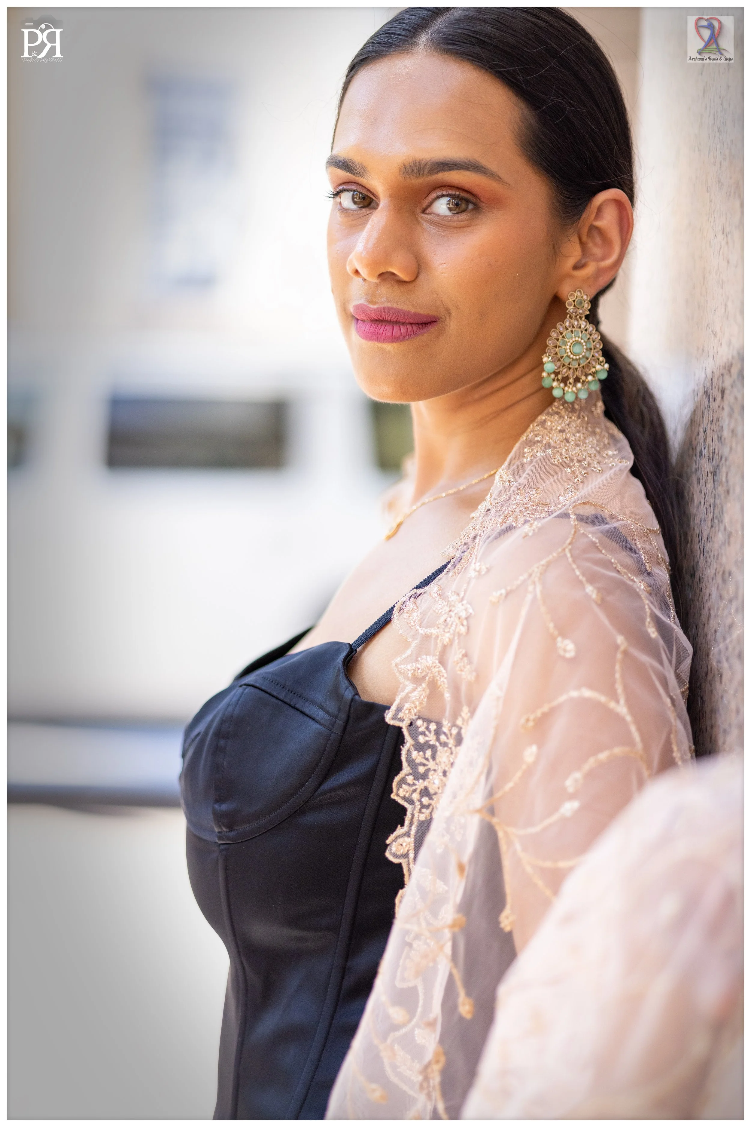 A woman with dark hair, wearing earrings and a pink lace shawl, leans against a wall and looks at the camera with a calm expression.