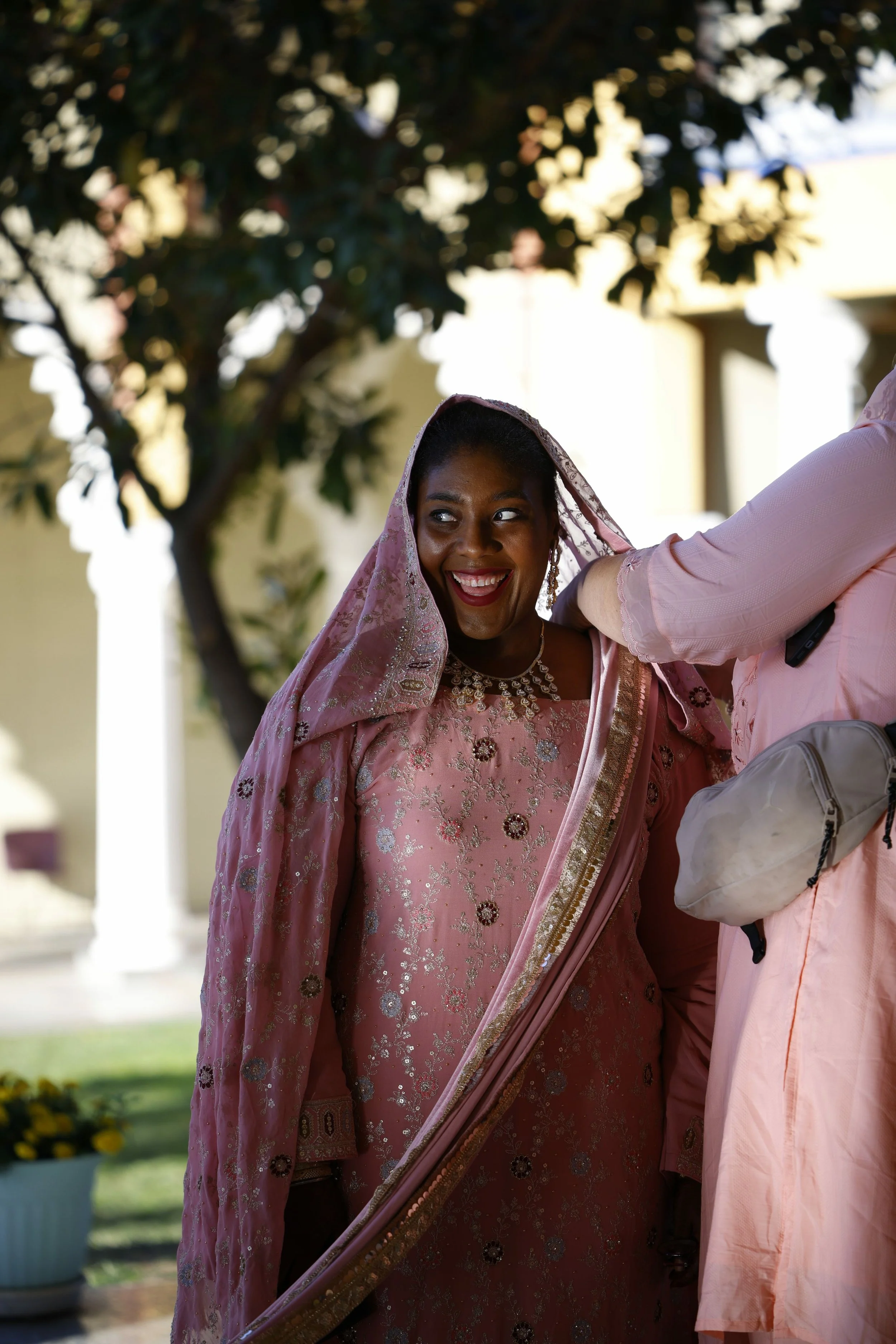 A woman in a traditional pink outfit with a matching veil, smiling and standing outdoors under a tree.
