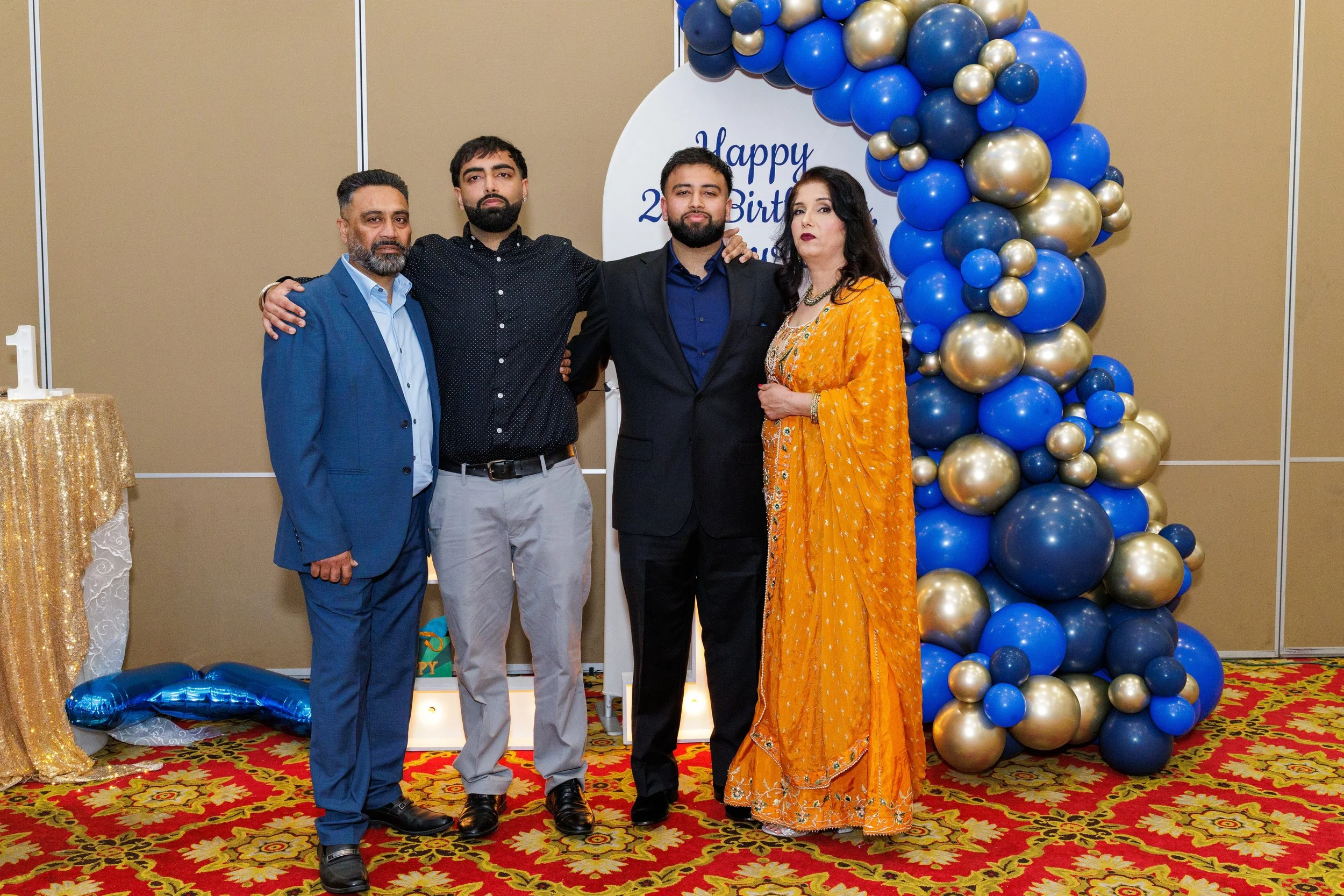 Family celebrating a birthday, with two men and a woman dressed formally, standing in front of a colorful balloon decoration and a 'Happy Birthday' sign, on a patterned carpet.