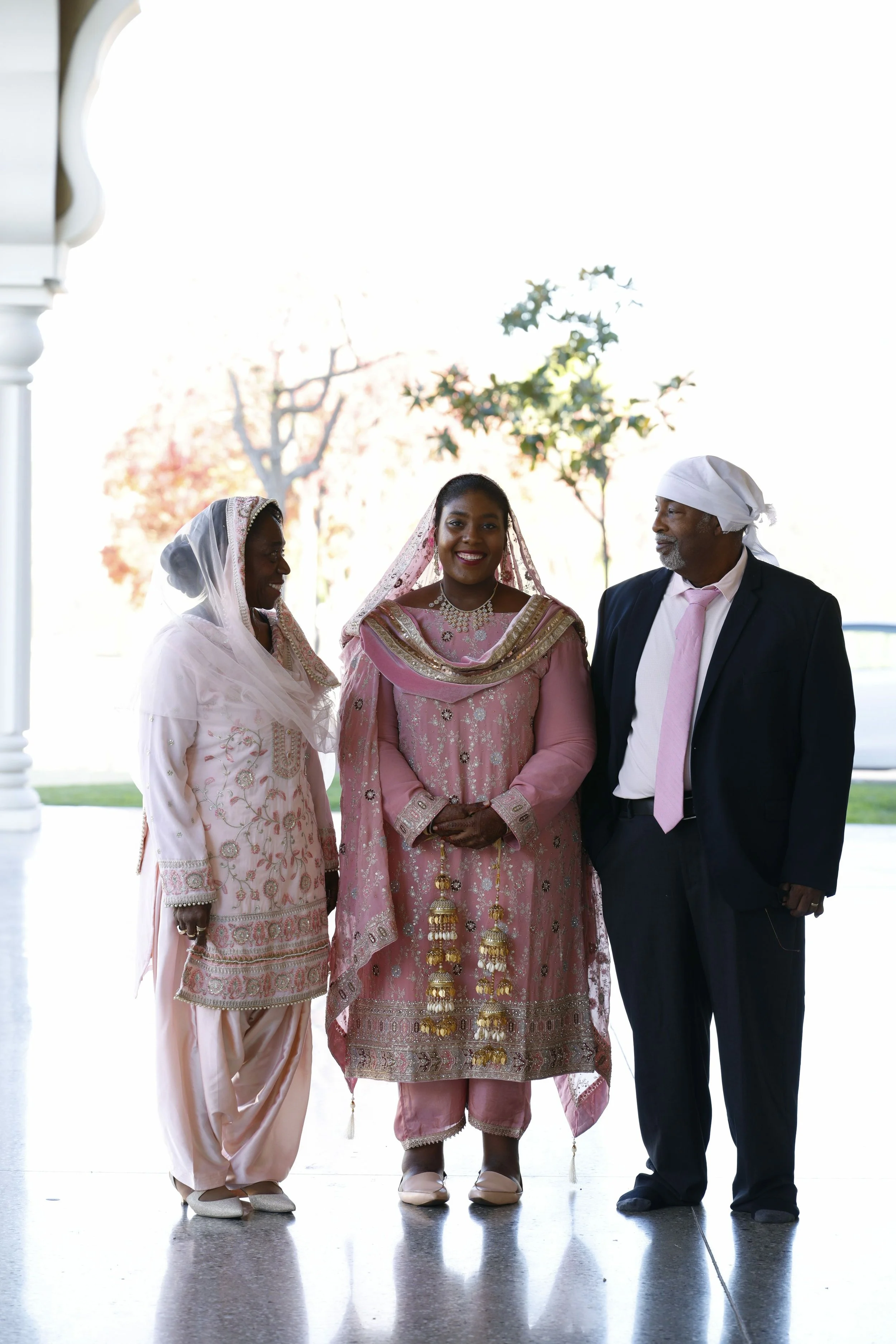 Three people at a wedding, an African woman in pink traditional attire, a woman in white traditional dress, and a man in a suit with a white head covering, all smiling.
