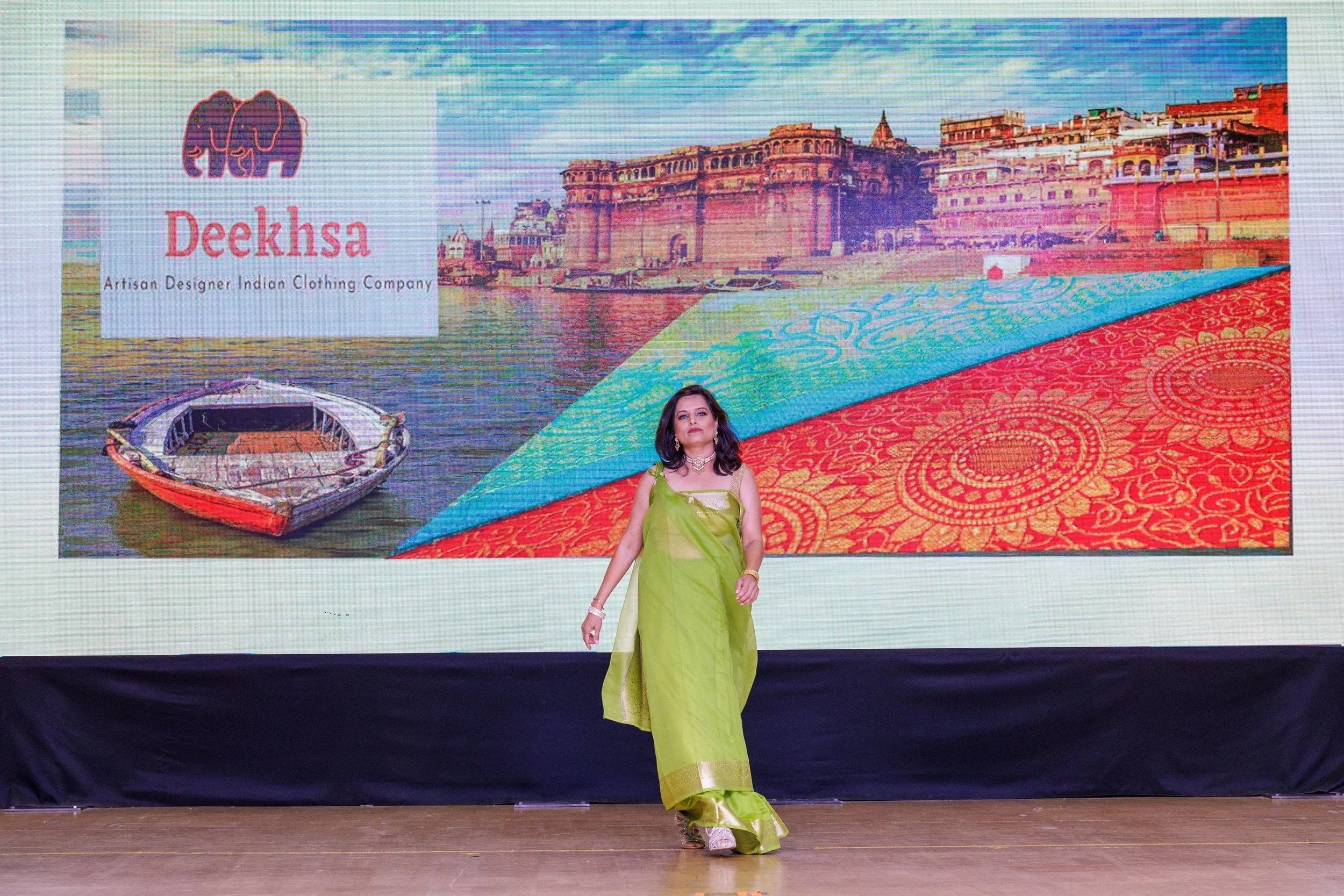 A woman in a bright green saree walks on a stage with a large colorful backdrop showing an image of a boat on a river, historic buildings, and a patterned textile. The backdrop features a logo and the text 'Deeksha Artisan Designer Indian Clothing Co