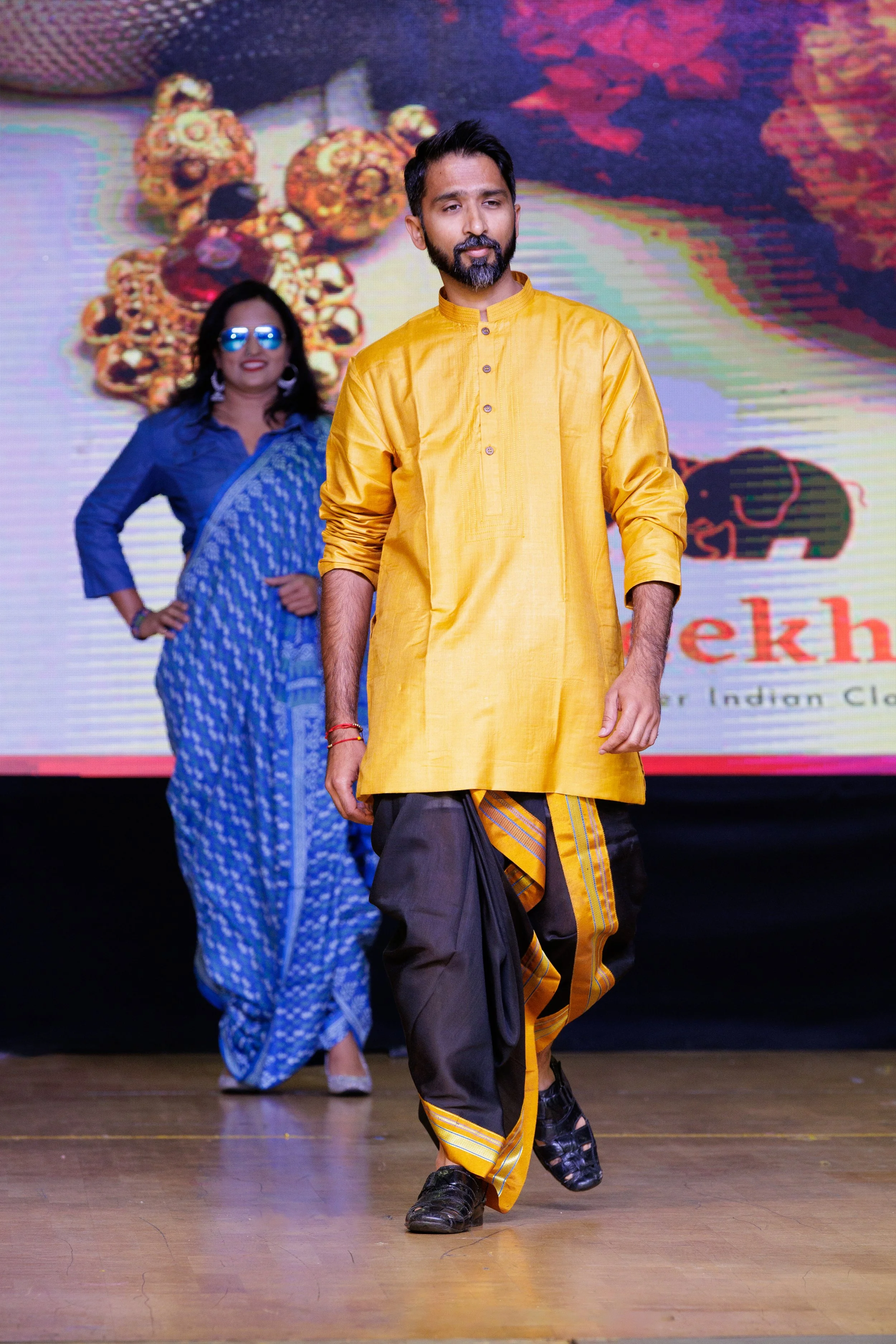 A man walking on stage wearing traditional Indian attire, with a woman in a blue sari behind him, during an event or fashion show.