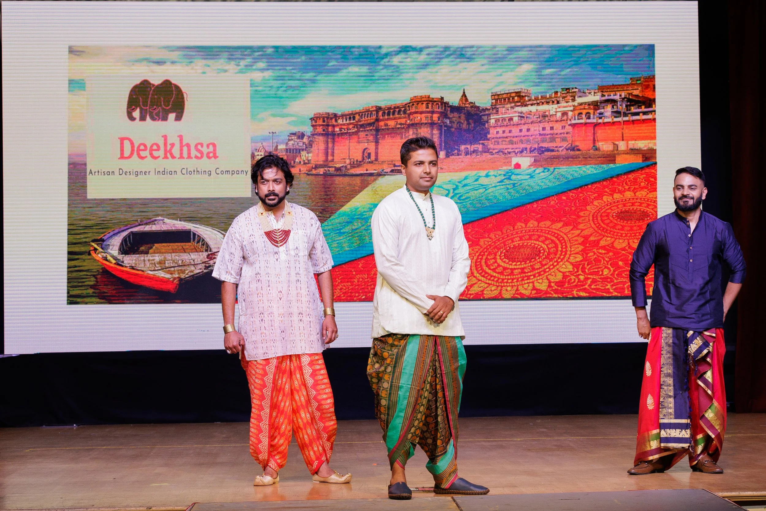 Three men standing on stage in traditional Indian clothing in front of a large screen with a colorful background and the text "Deeksha" and "Artisan Designer Indian Clothing Company".