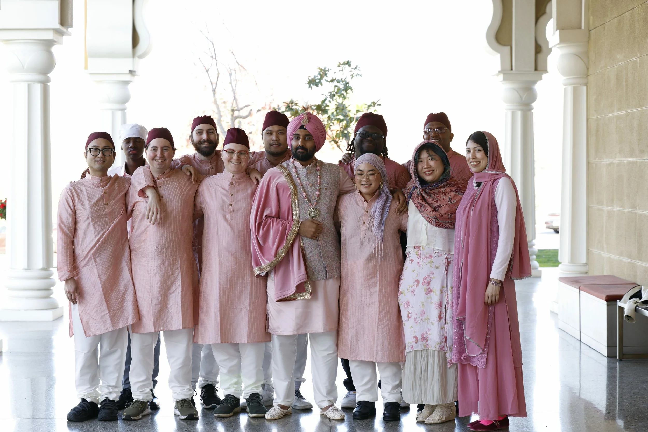 A diverse group of people dressed in traditional Indian clothing, posing together outside a building with columns. Some are wearing pink and white outfits, and one man is dressed in more ornate attire with jewelry and a pink turban.