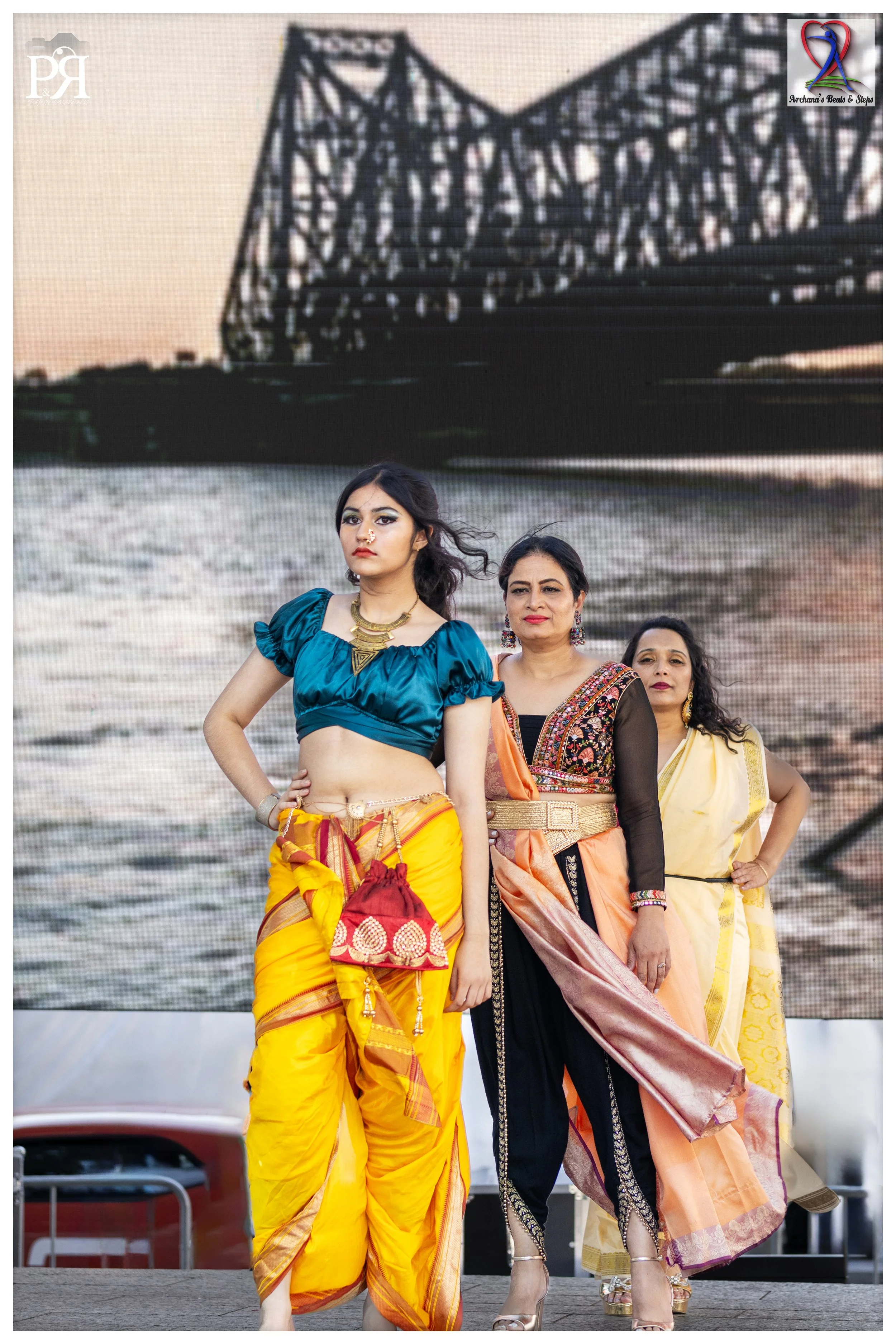 Three women in traditional Indian attire standing outdoors in front of a large digital screen displaying a modern building with a river in the foreground during sunset.
