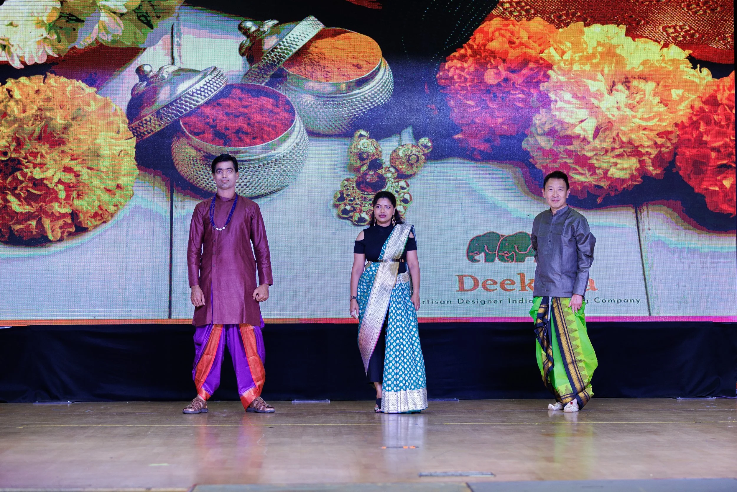 Three people standing on a stage, dressed in traditional Indian attire, with a large digital screen in the background displaying colorful decorations and text that reads 'Deeksha'.