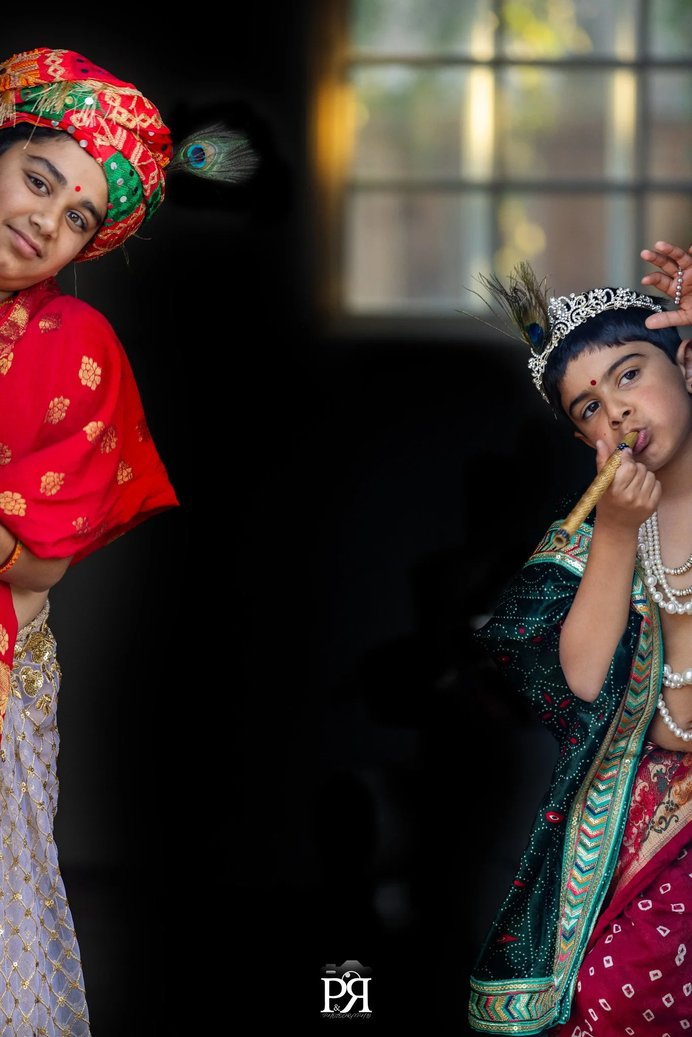 Two children dressed in traditional Indian costumes, one girl on the left wearing a red saree and colorful turban, and a boy on the right wearing a king or prince costume with a crown and playing a themed flute, standing indoors with a window in the 