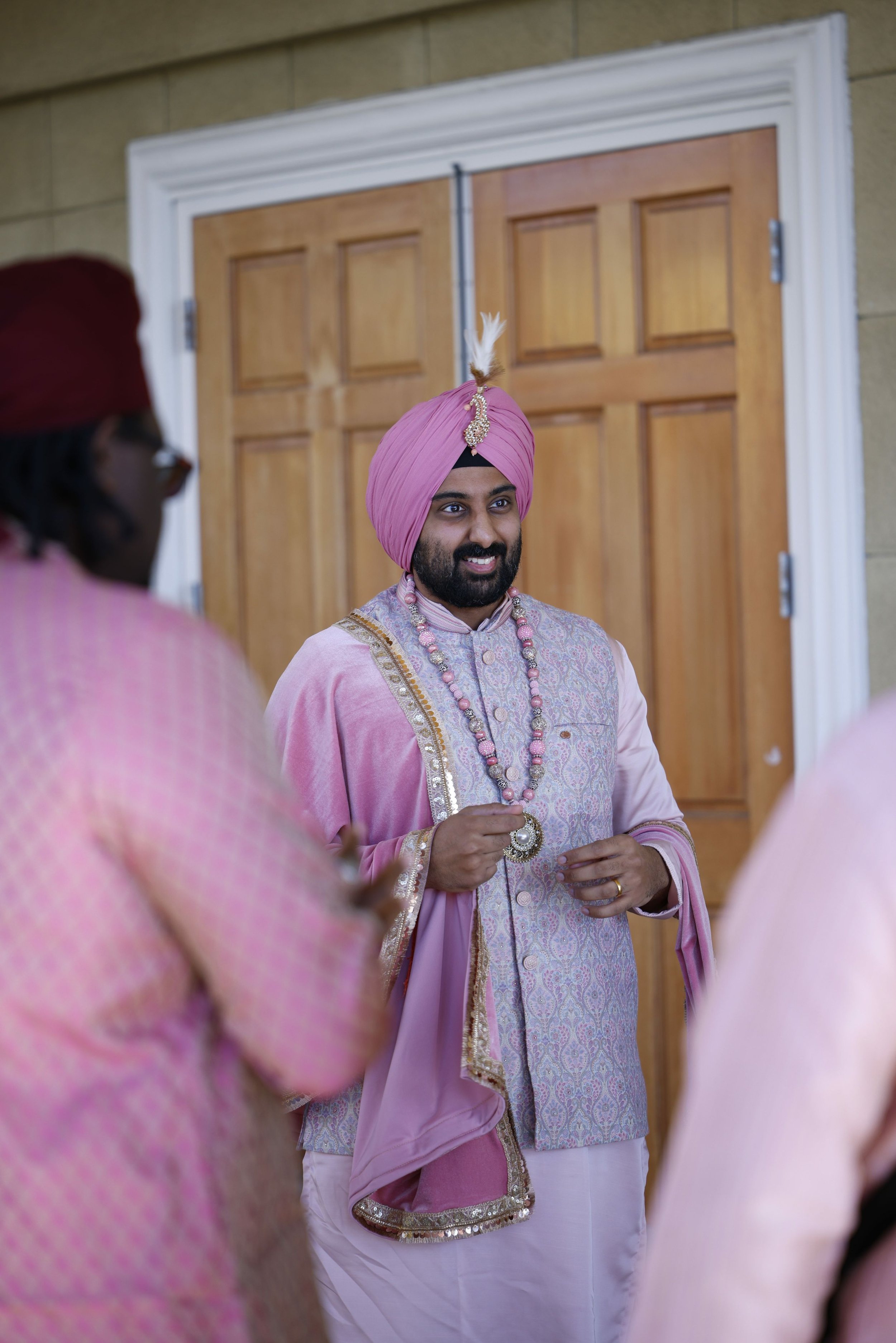 A man dressed in traditional Indian attire, wearing a pink turban with a feather and jewelry, is smiling at an Indian wedding ceremony.