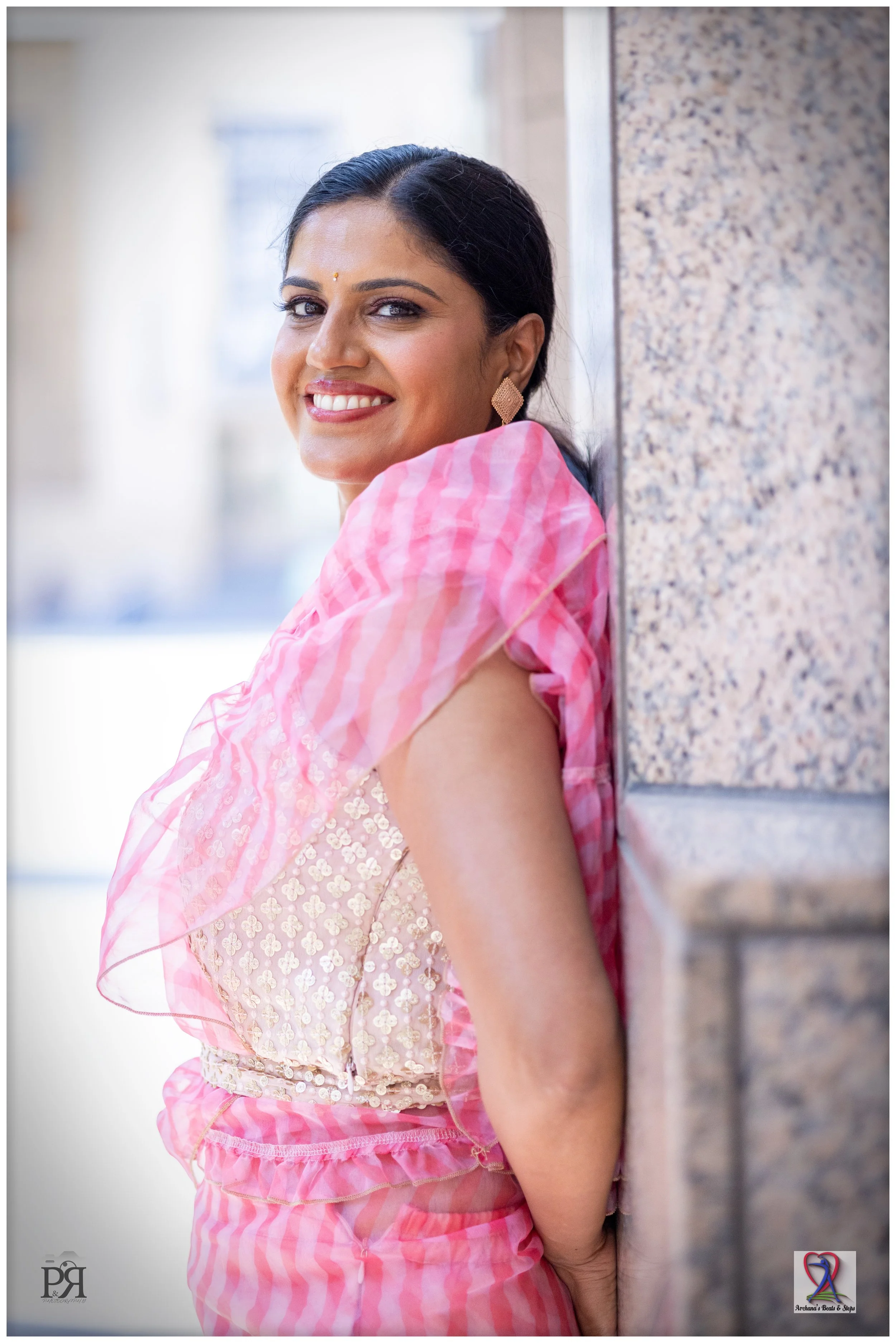 A woman dressed in traditional Indian attire, leaning against a stone wall, smiling at the camera, with dark hair tied back, wearing earrings, and a pink sari with decorative embellishments.
