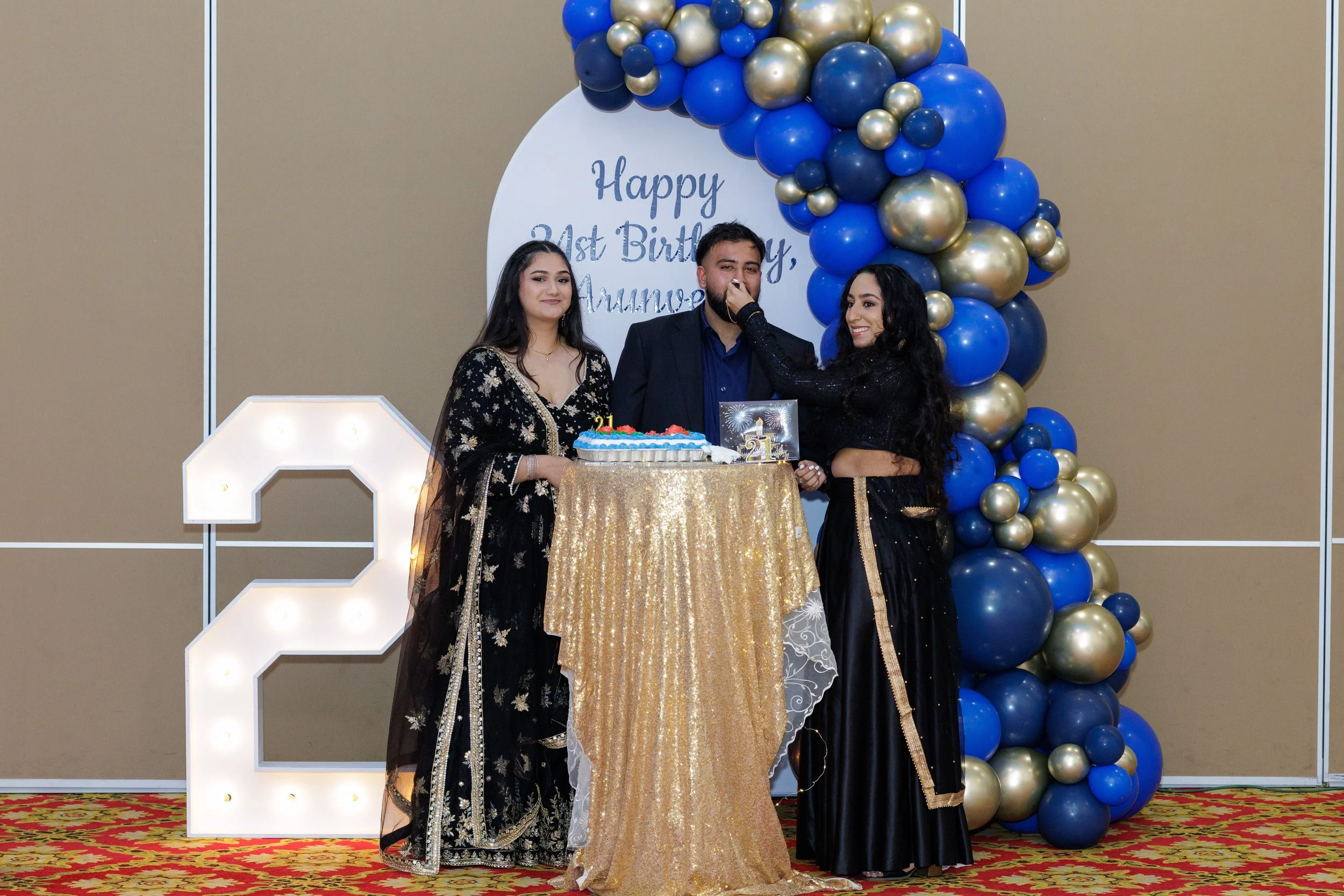 Three people celebrating a birthday at a decorated event, with a large illuminated number '2', a gold tablecloth, a birthday cake, and balloon decorations in blue, gold, and silver, with a sign that reads 'Happy 21st Birthday, Amna' in the background