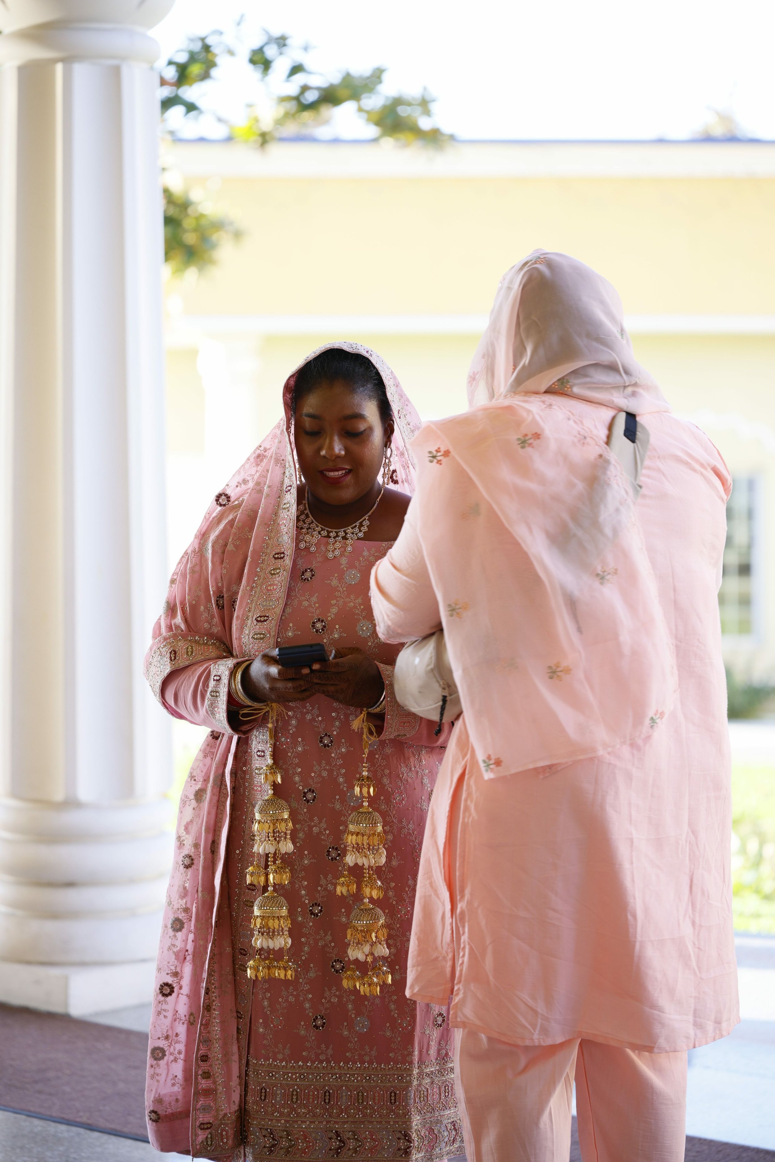 Two women in traditional Indian attire, one with a pink embroidered dress and the other with a light pink shawl over her head, standing outdoors, looking at a cellphone.